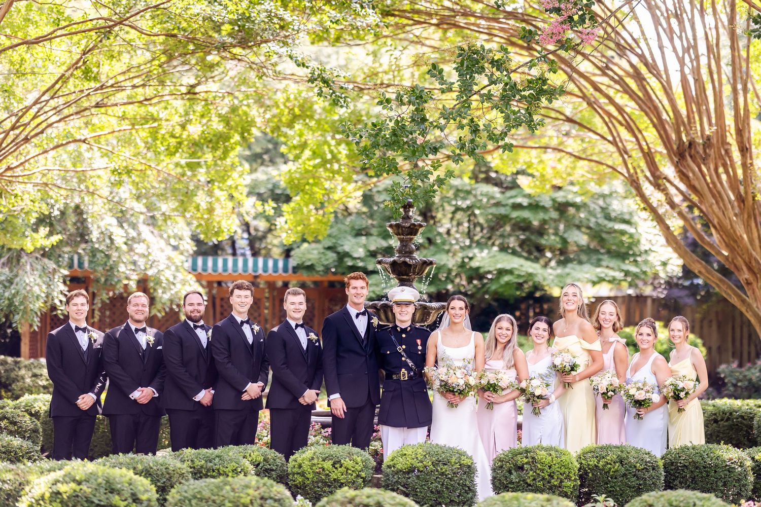 Wedding party poses for formal group photos under trees in an outdoor garden setting.