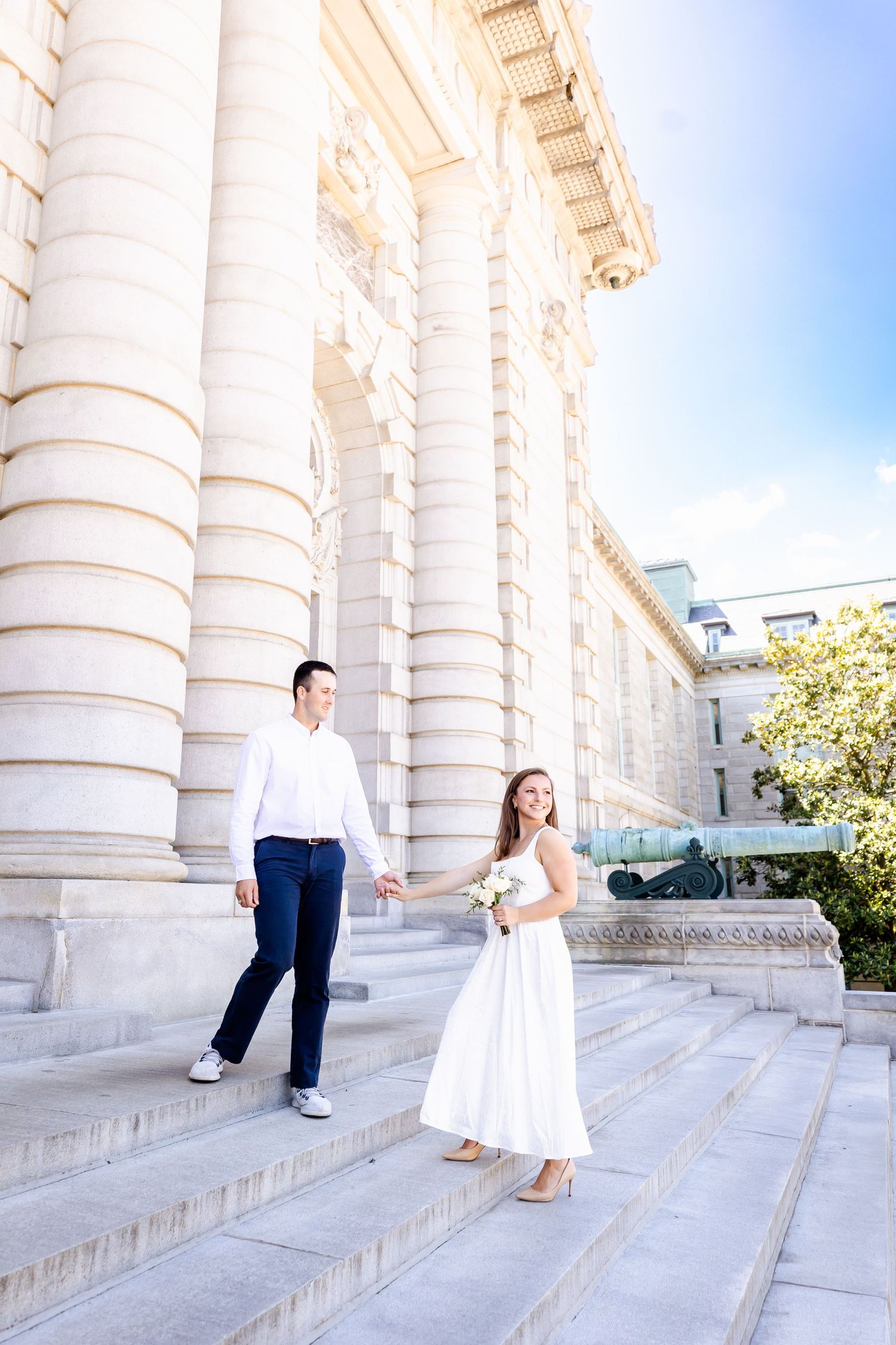 A couple in formal attire poses playfully on white marble steps between classical columns on a sunny day.
