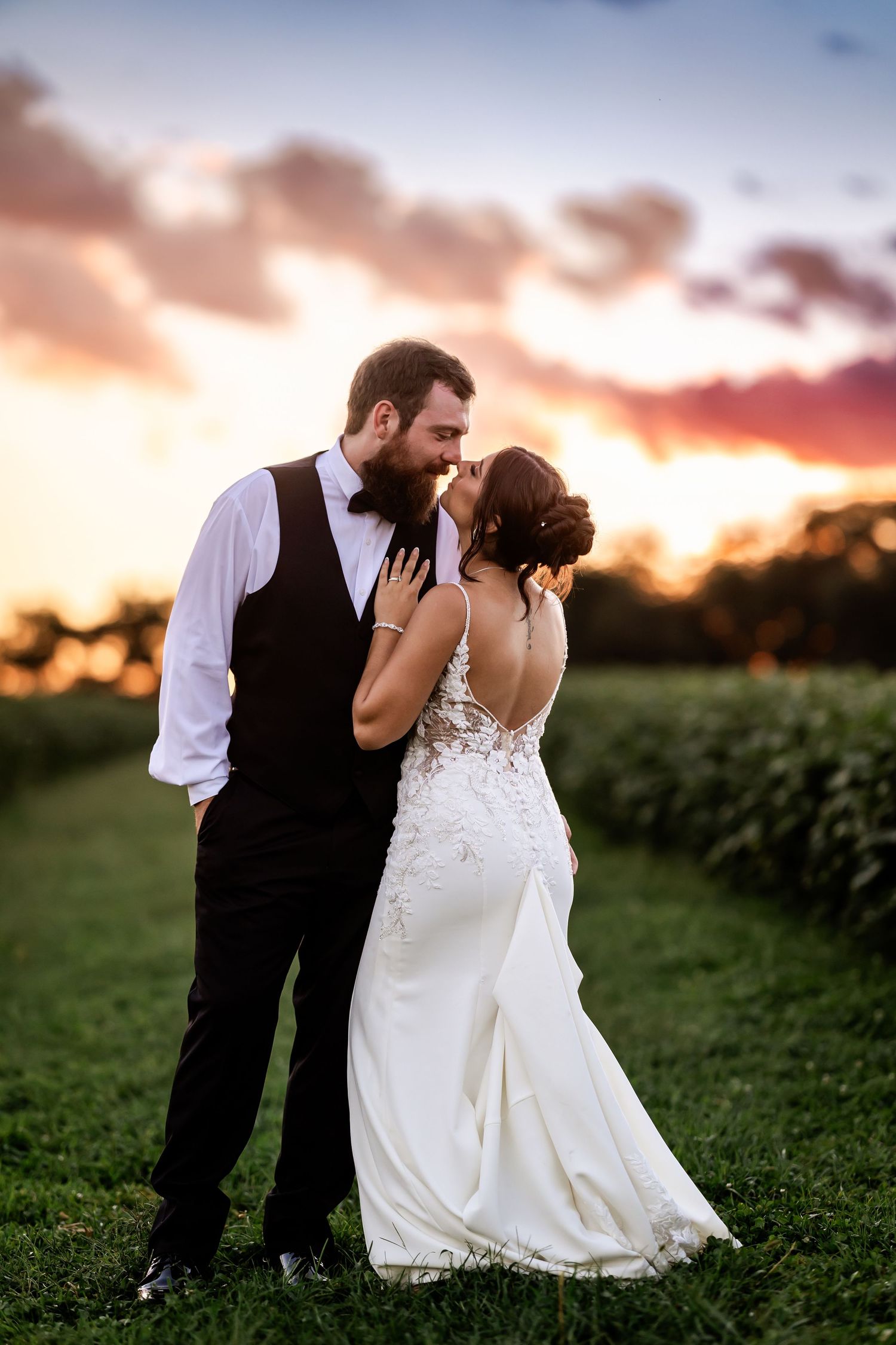 Bride and groom share a romantic almost-kiss at sunset in a lush green field, wearing elegant wedding attire.