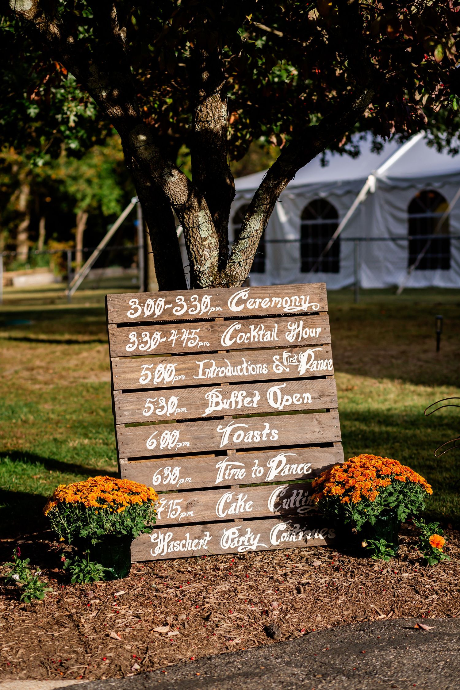 Wooden wedding schedule signs display event timeline details next to orange flowers in a garden setting.