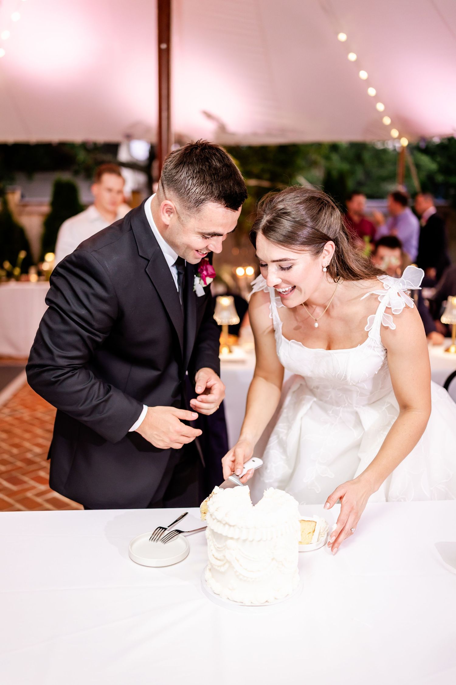 Newlyweds cut their white wedding cake together under a reception tent with string lights.
