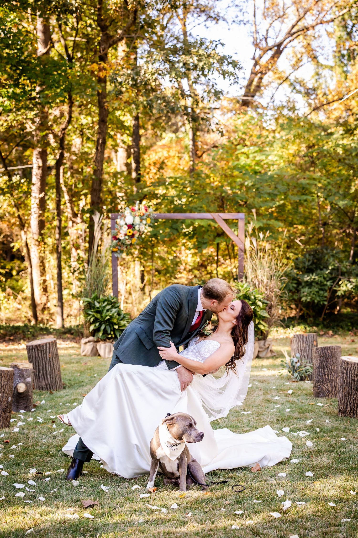 Romantic wedding couple shares intimate kisses on grass in a sunlit forest setting with scattered flower petals.