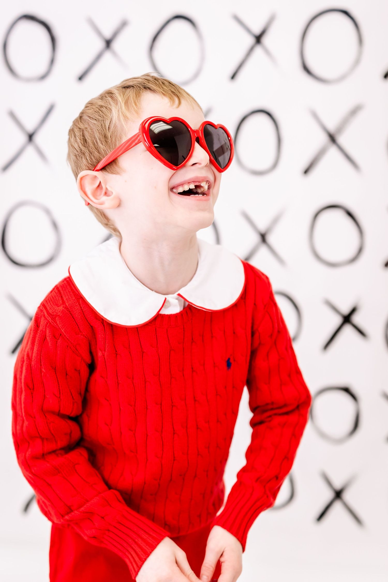 Playful child in red sweater wearing heart-shaped red sunglasses poses against a wall decorated with tic-tac-toe design.