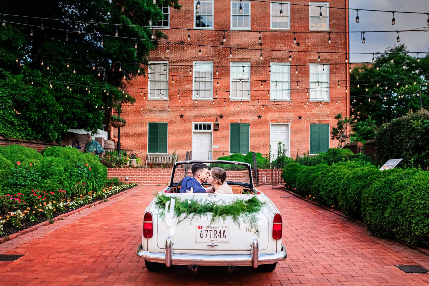 A white classic convertible car parked in front of a red brick building with couple seated in the backseat.