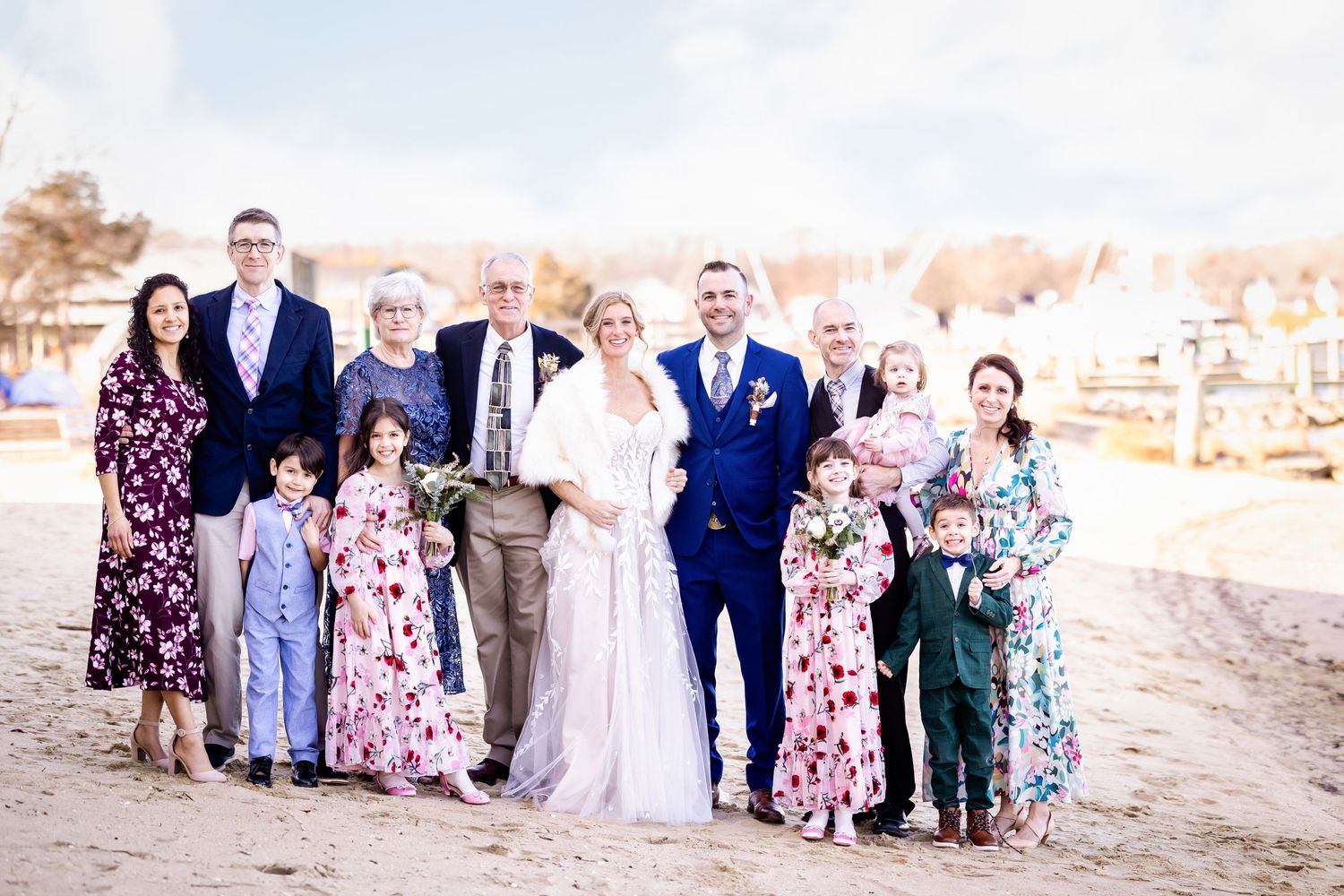 A wedding party poses together on a beach at sunset wearing floral dresses and formal attire.