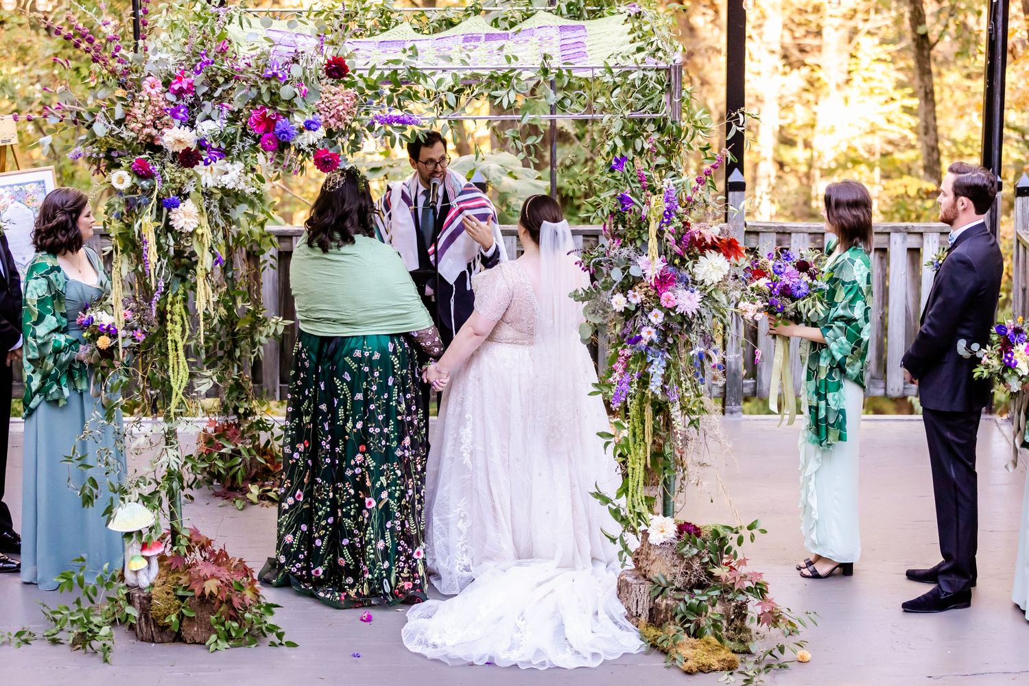 Wedding ceremony under a floral arch with purple and pink blooms as guests look on.