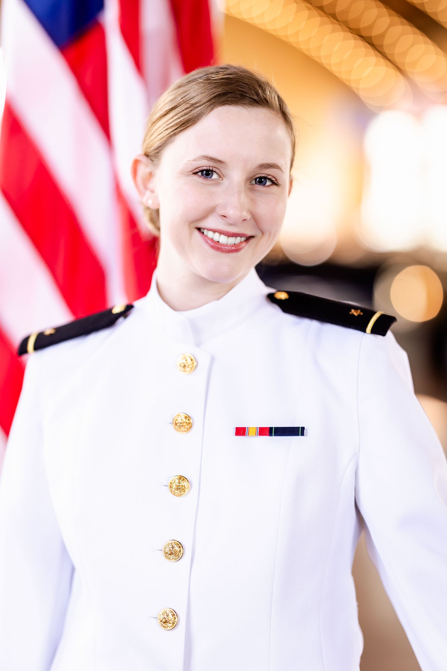 A service member in dress whites poses in front of an American flag.