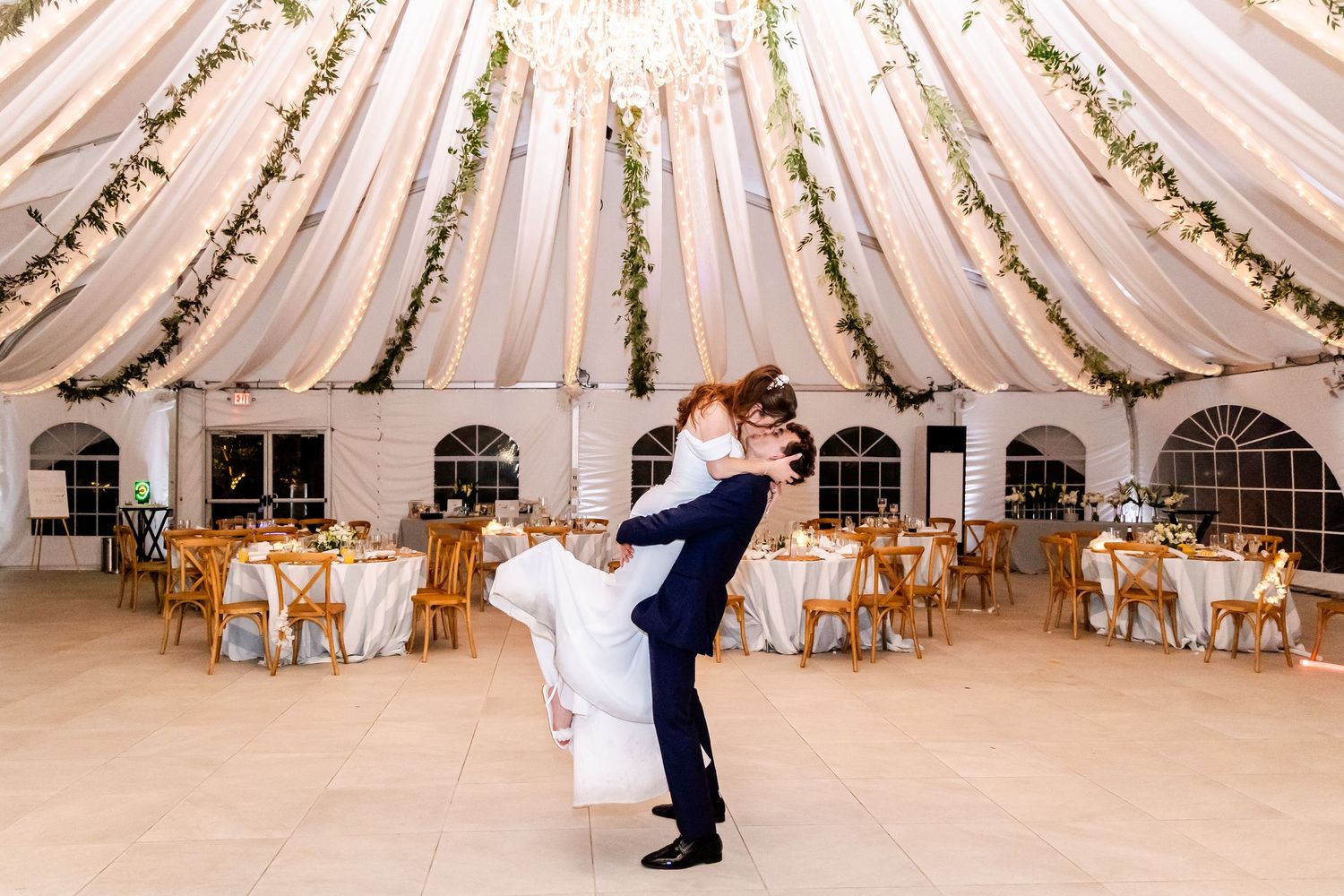 Elegant wedding tent interior with white draped fabric ceiling and wooden cross-back chairs.