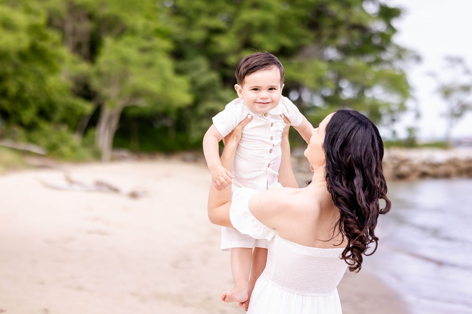 A parent and young child share a tender moment on a beach with trees and water in the background.