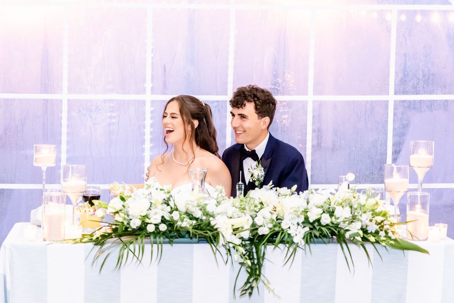 White floral arrangements and greenery decorate a wedding reception head table with sheer backdrop.
