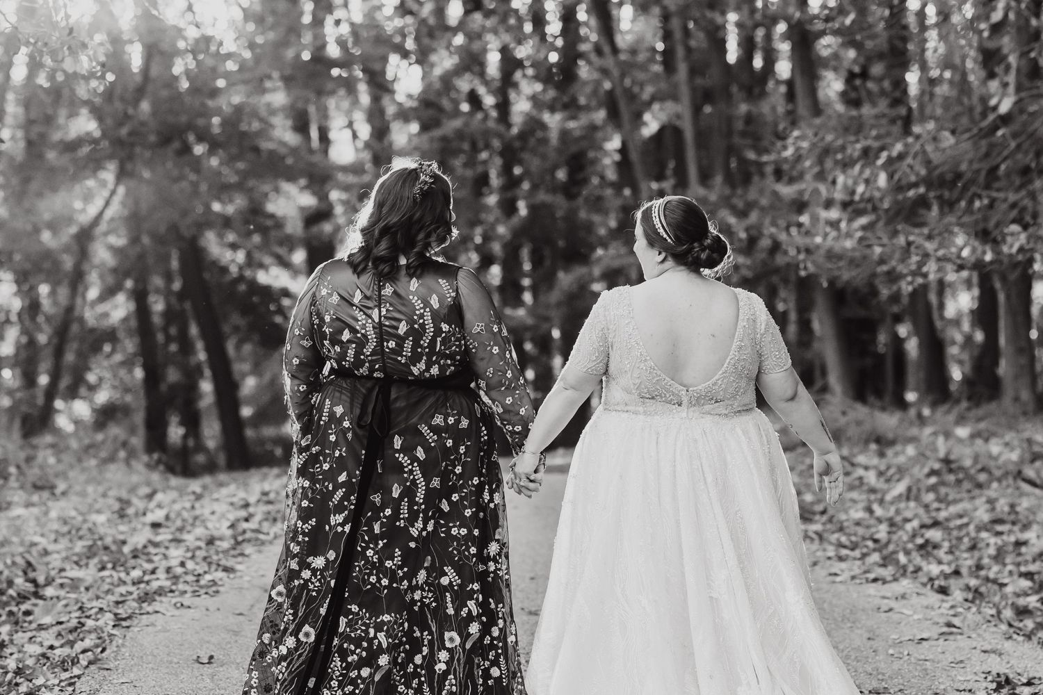 Two figures walking down a wooded path wearing flowing floral and white dresses in black and white photography.