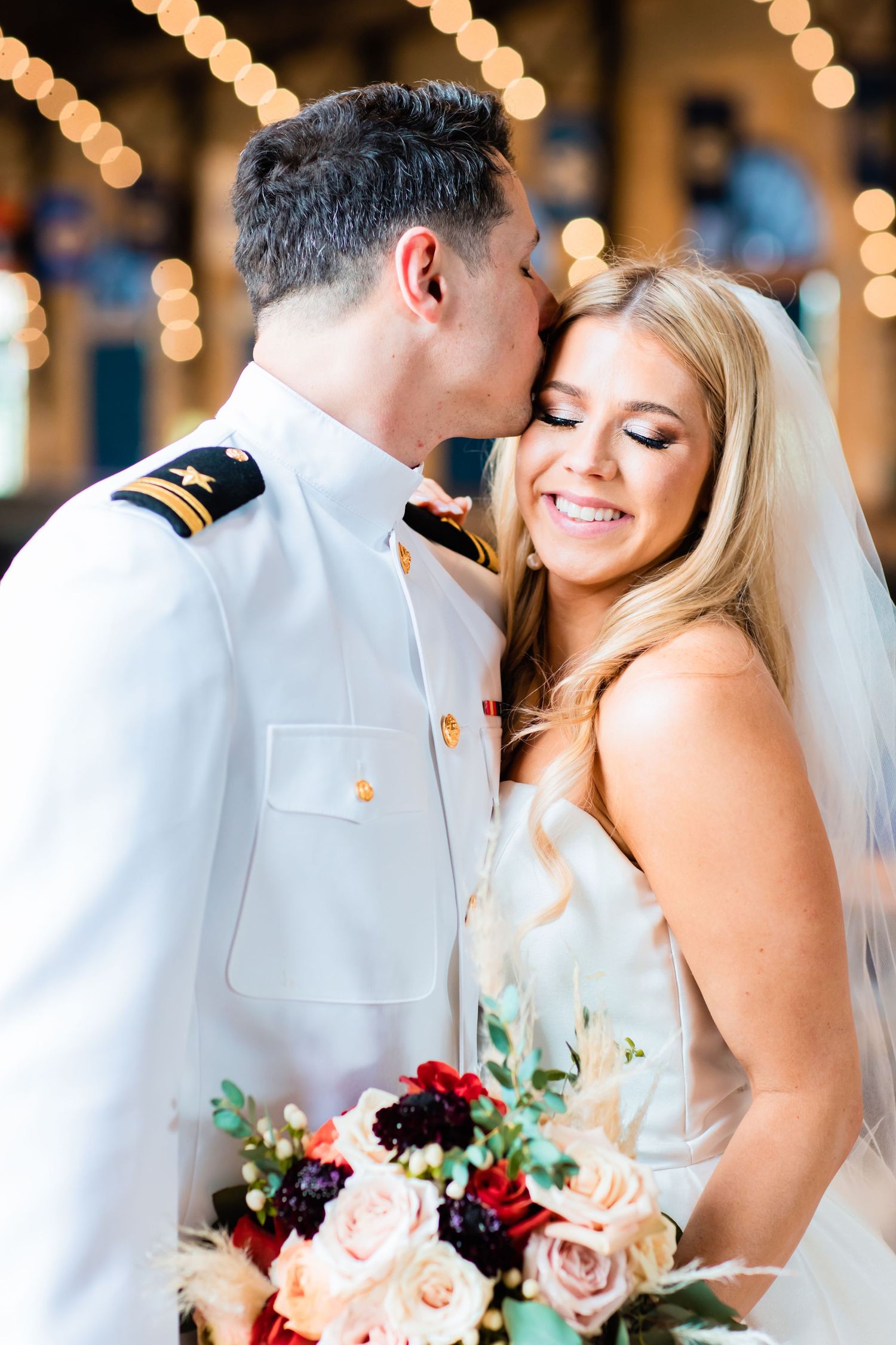 A military officer in dress whites embraces his bride who holds a pink and burgundy bouquet under romantic string lights.