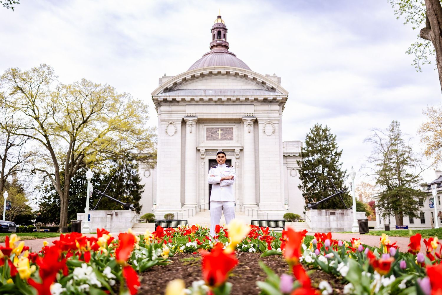 A white classical gazebo structure with red tulips blooming in the foreground garden at a park on a spring day.