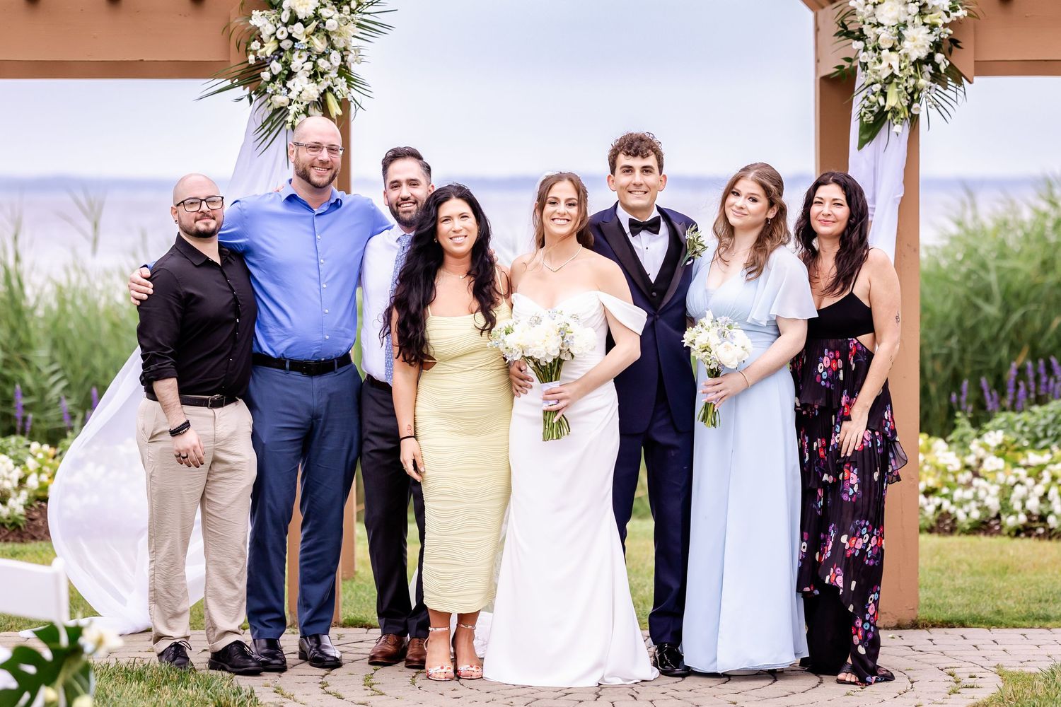 Large wedding party poses for a group photo under a wooden pergola decorated with white floral arrangements.