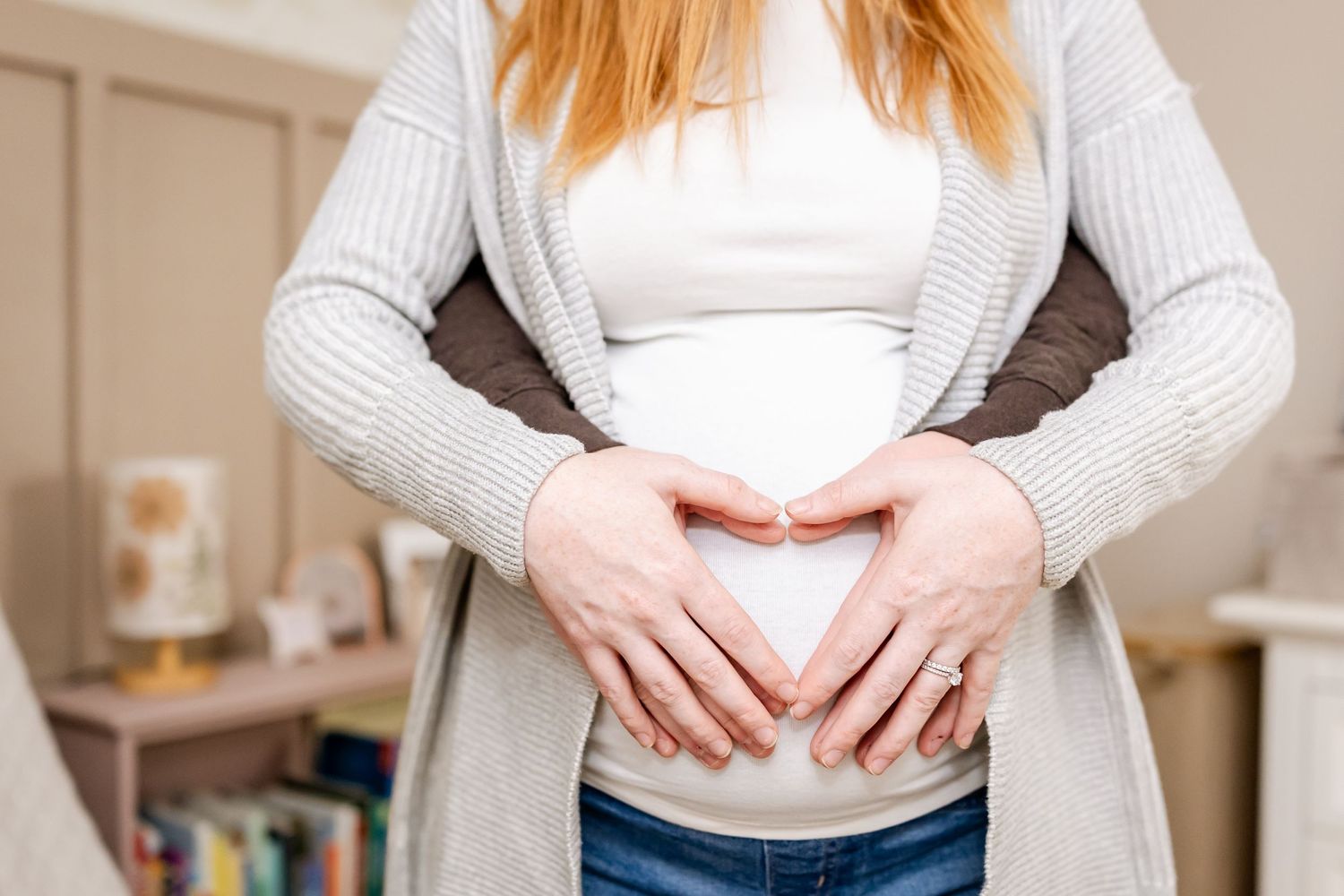 Hands forming heart shape over pregnant belly while wearing white sweater and jeans in home setting.