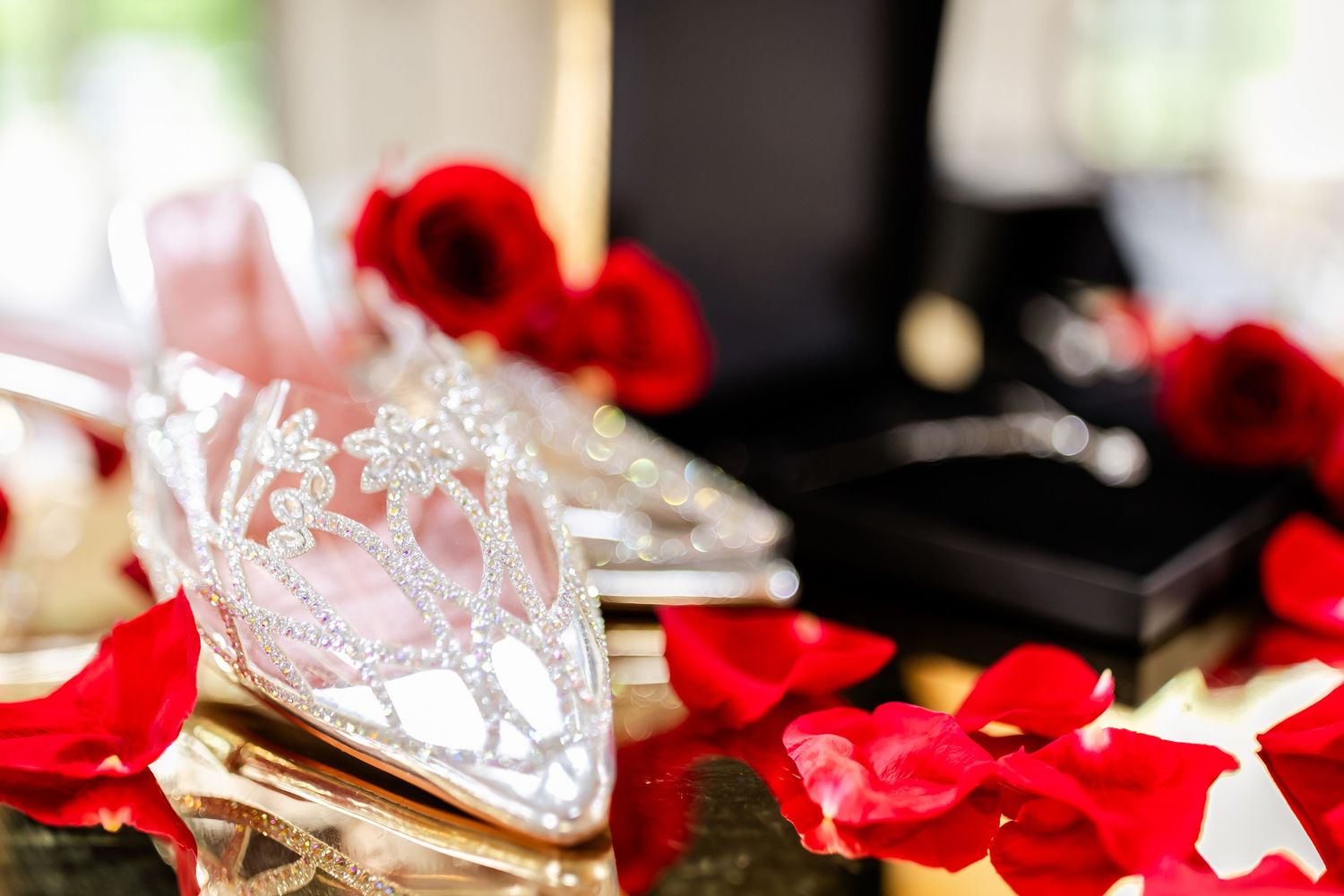 Crystal tiara and red rose petals arranged on reflective black surface for wedding decoration.