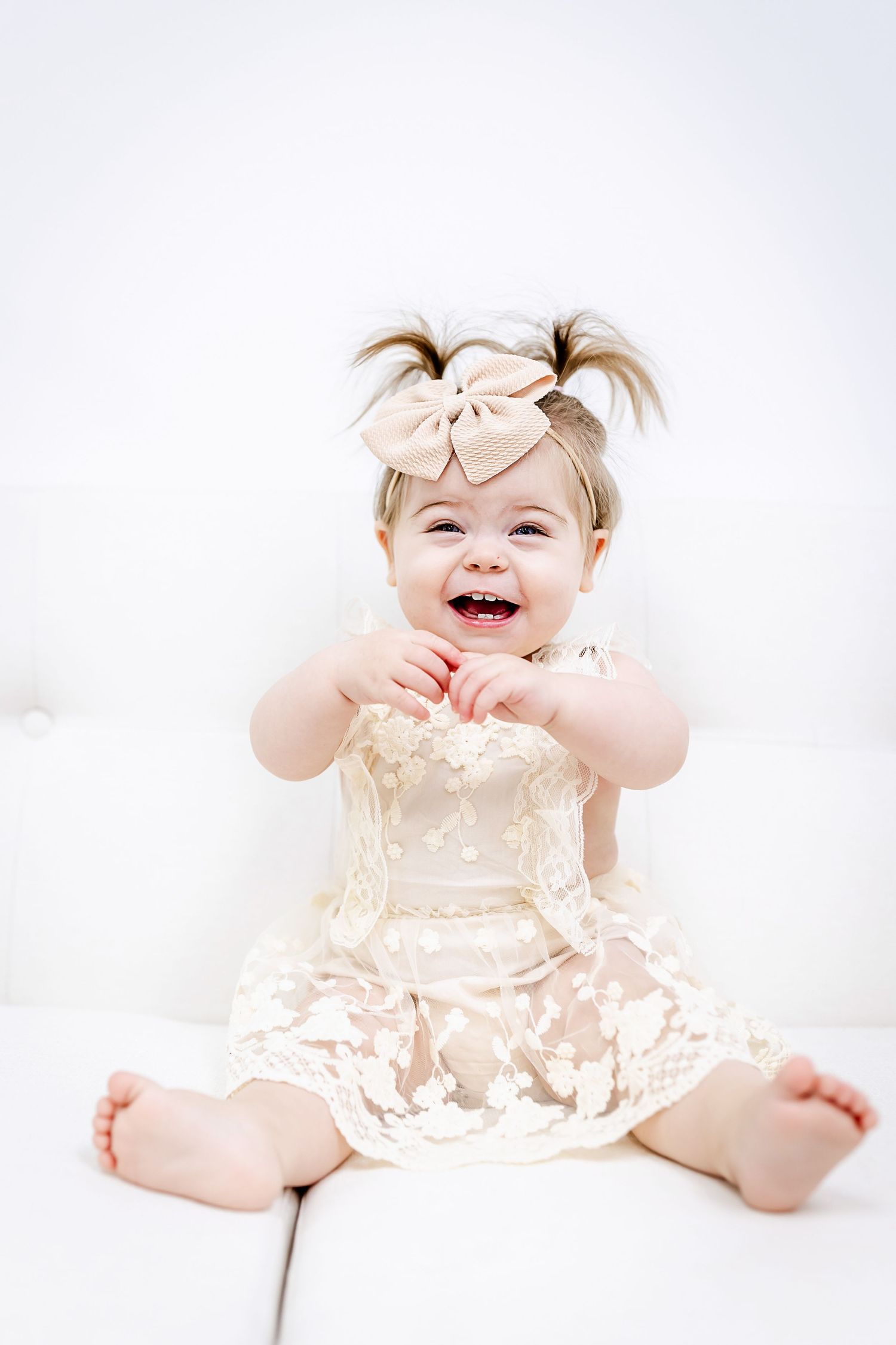 A baby in cream dress and bow laughs joyfully against a white background.