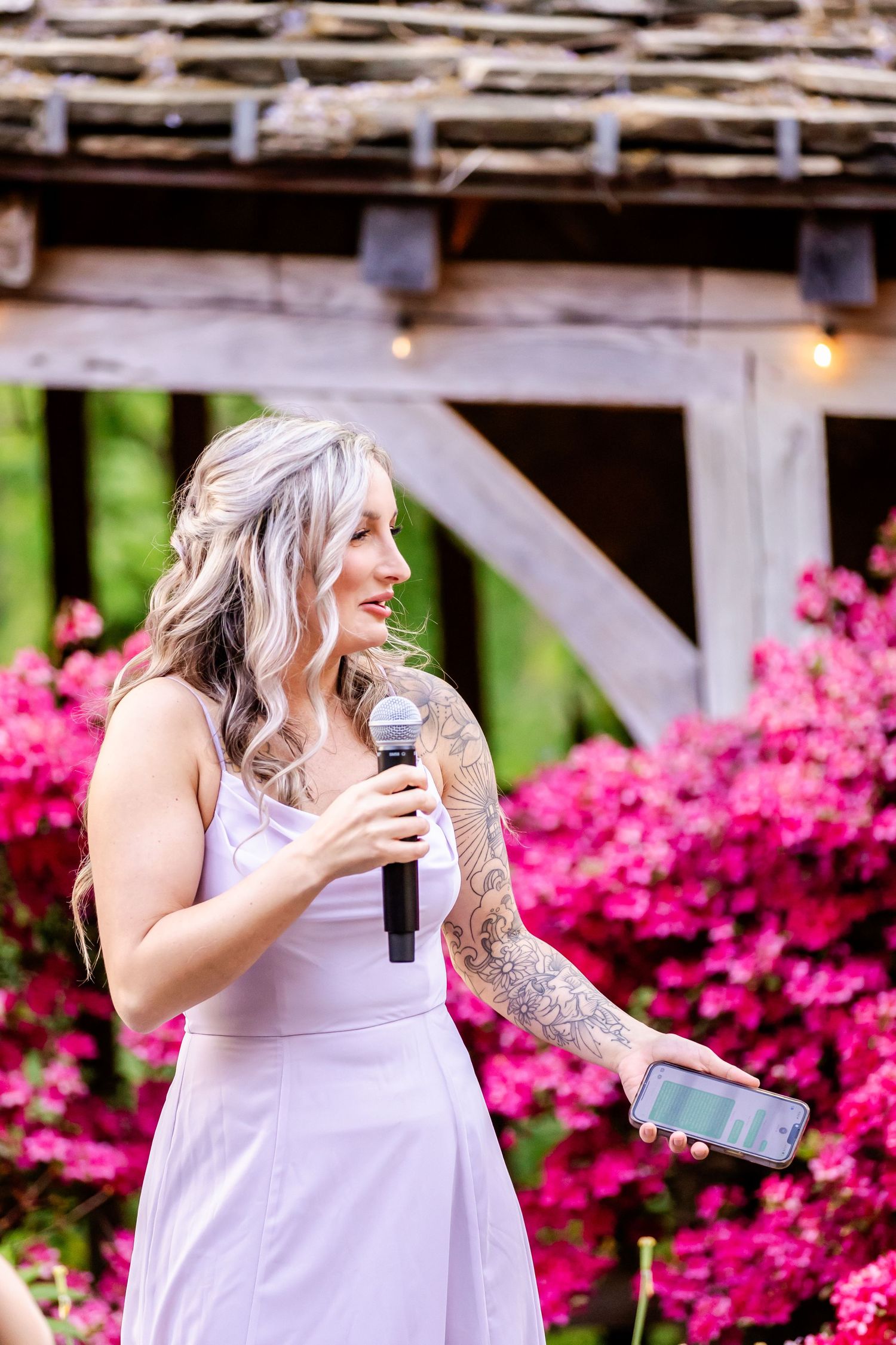 Person in purple dress gives speech with microphone at outdoor wedding venue with pink flowers.