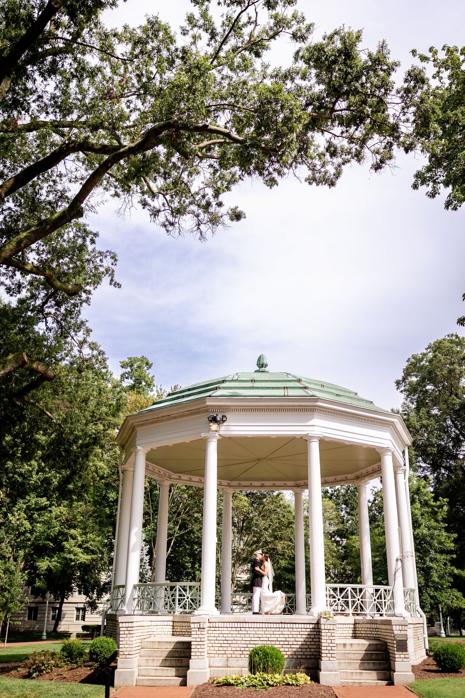 White gazebo with elegant columns surrounded by lush trees in a garden setting.