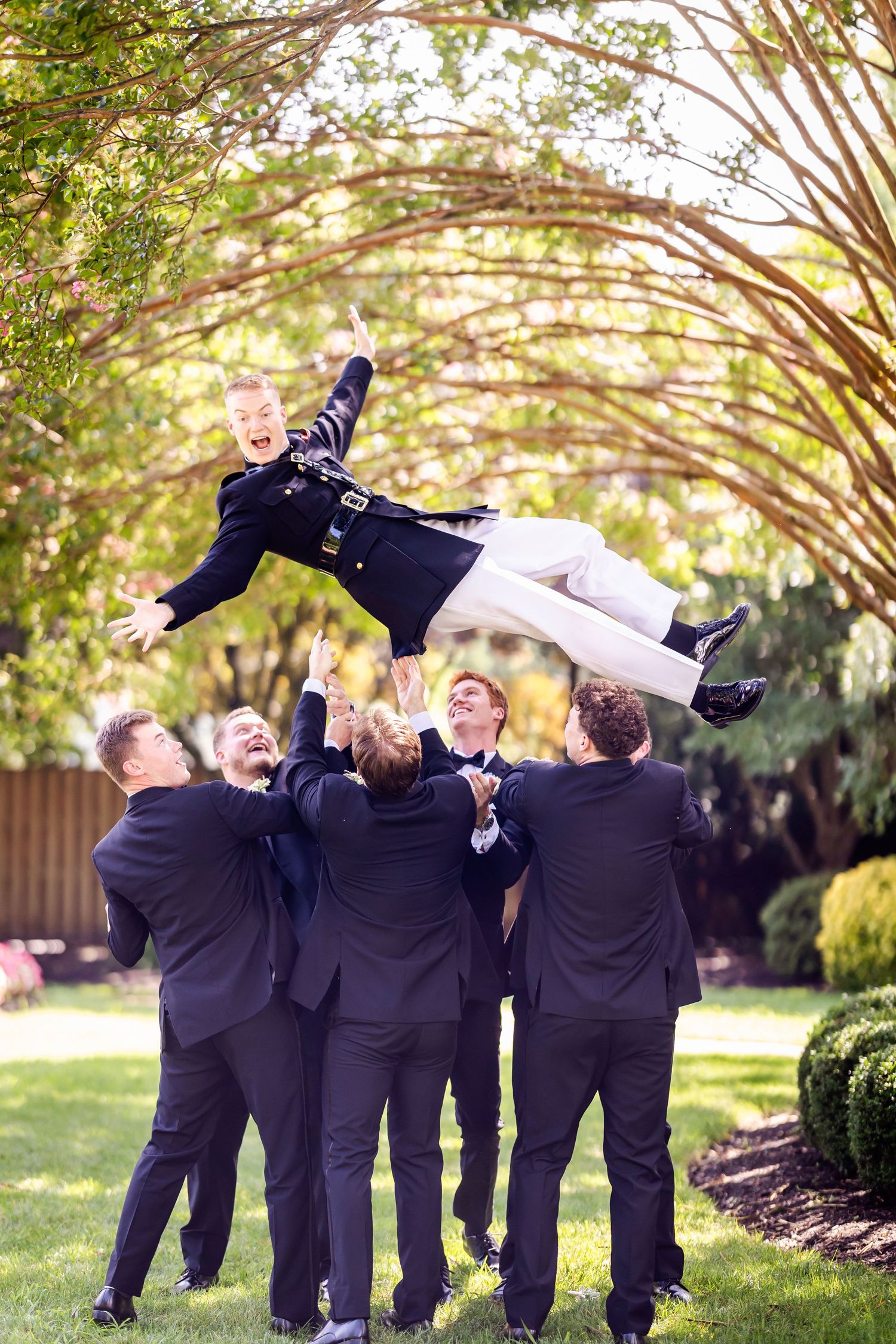 Groomsmen in navy suits playfully toss groom in air during outdoor wedding photo session.