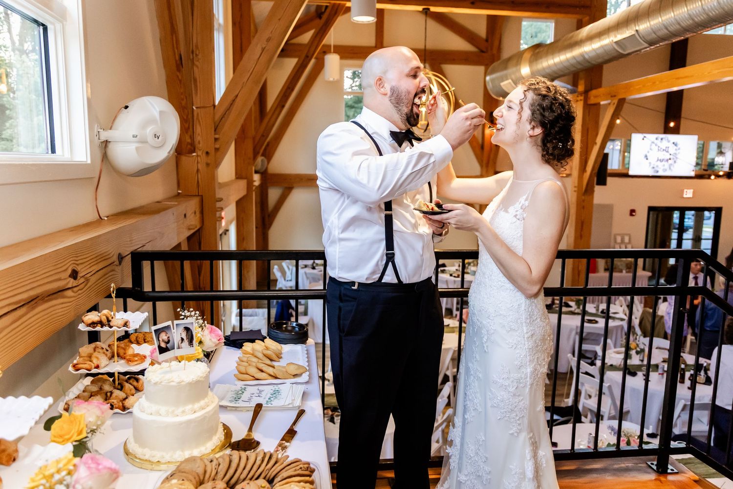 A wedding couple and guests gather around a rustic dessert table with pies and treats at a barn reception venue.