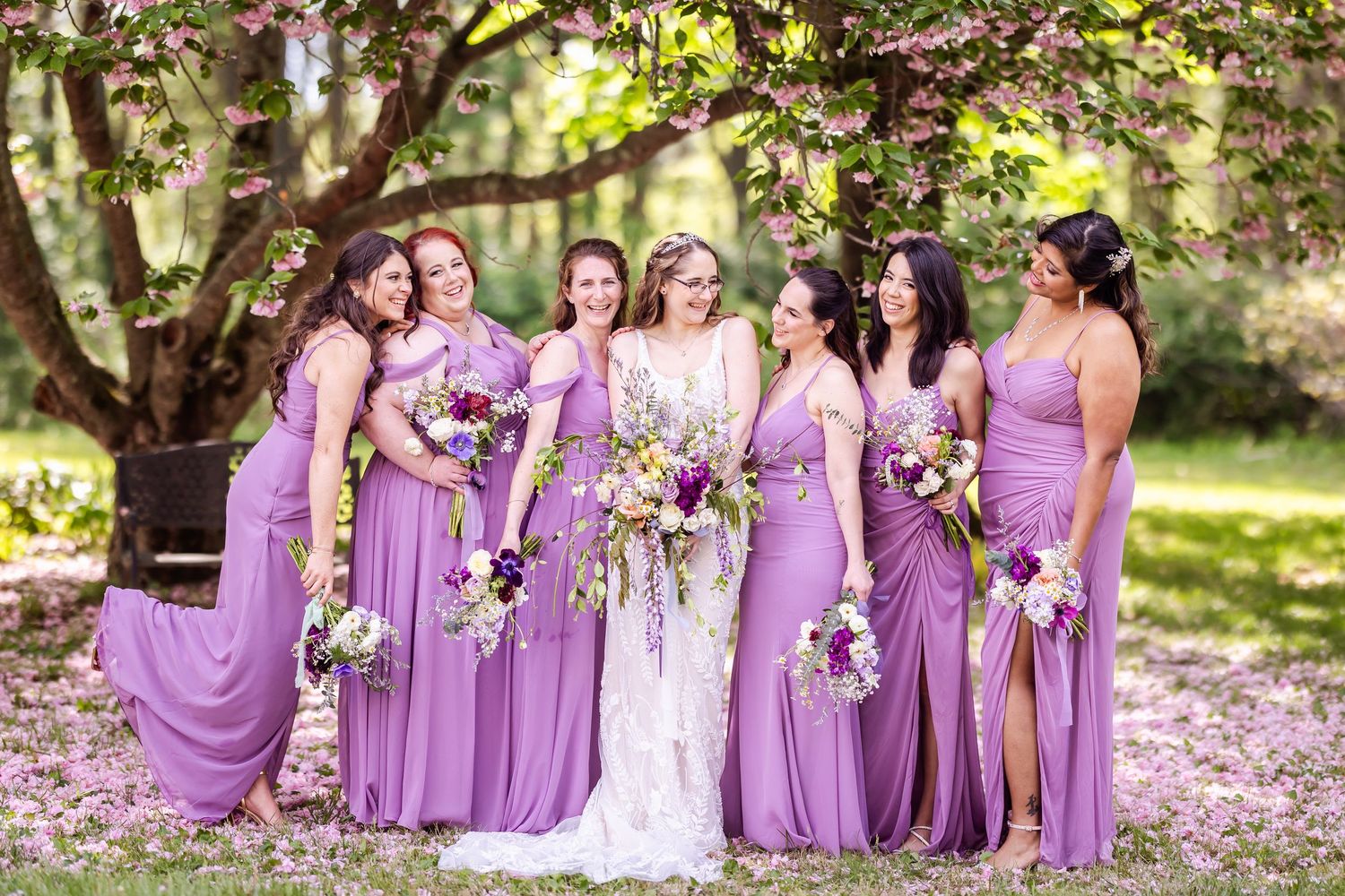 Wedding party wearing purple bridesmaid dresses poses together in a garden setting with blooming flowers.