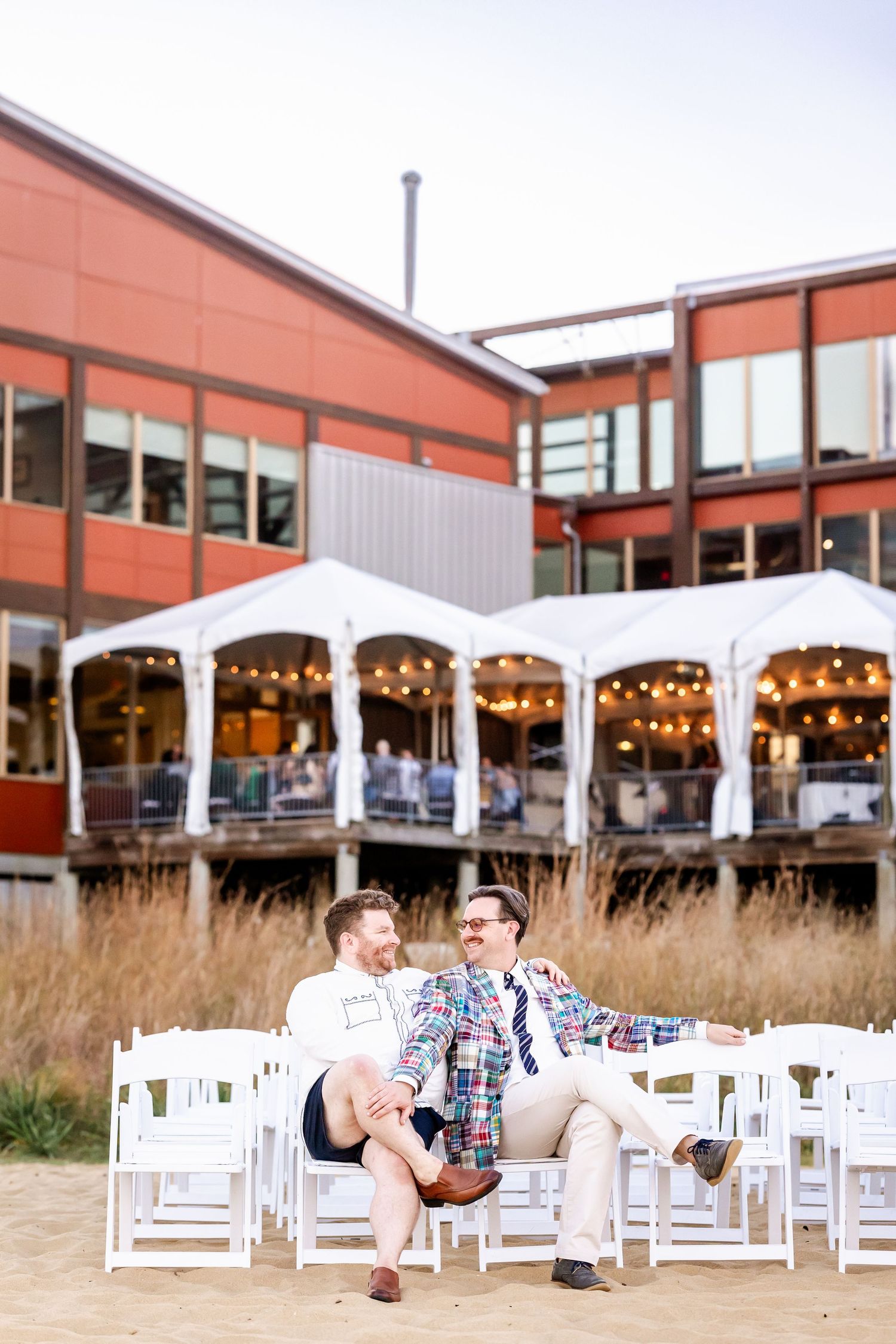 Couple relaxes in white Adirondack chairs on a wooden deck with string lights and tall grass at sunset.