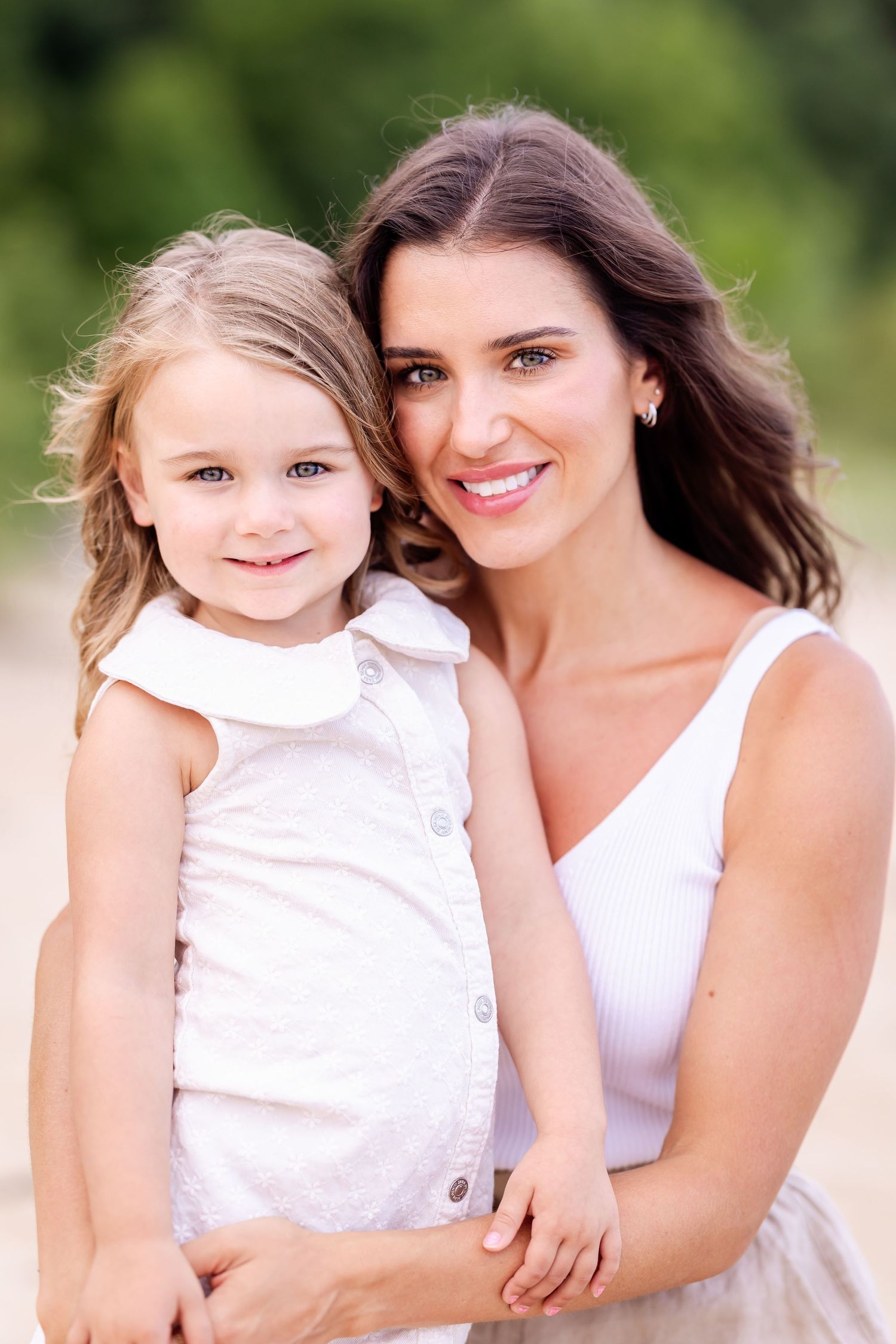 A mother and young daughter wearing matching white dresses pose together outdoors with a blurred green background.