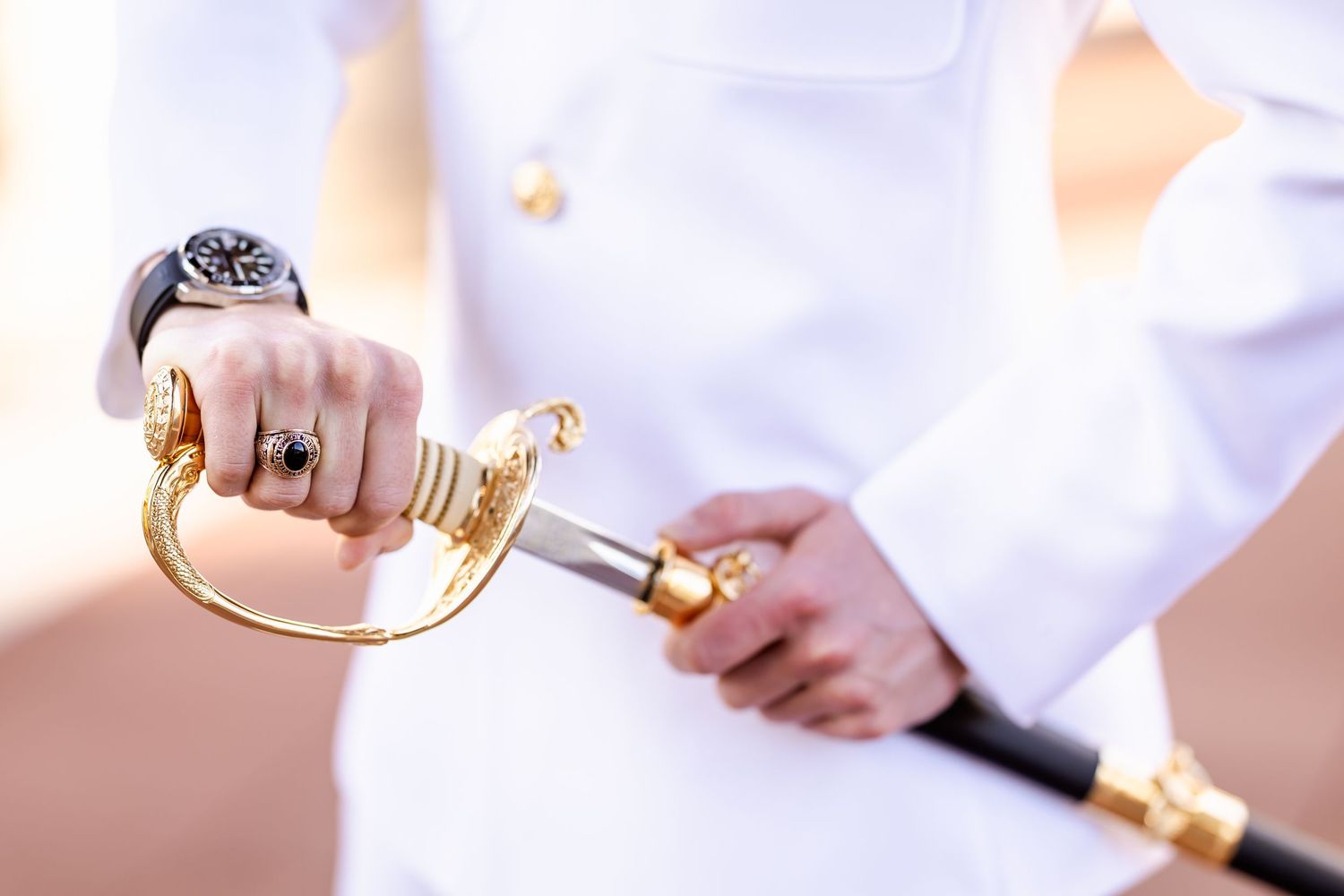 Military officer in white uniform holding a gold-handled ceremonial sword, wearing a ring and watch.