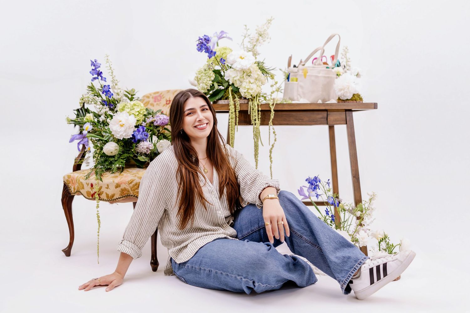 Young woman sitting on floor surrounded by spring flowers and floral arrangements on vintage furniture against white background.