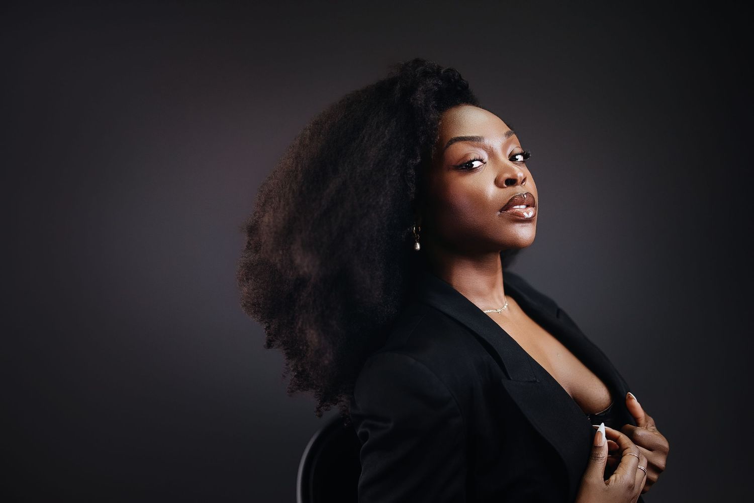 A confident Black woman with voluminous curly hair poses in a black blazer against a dark studio background.
