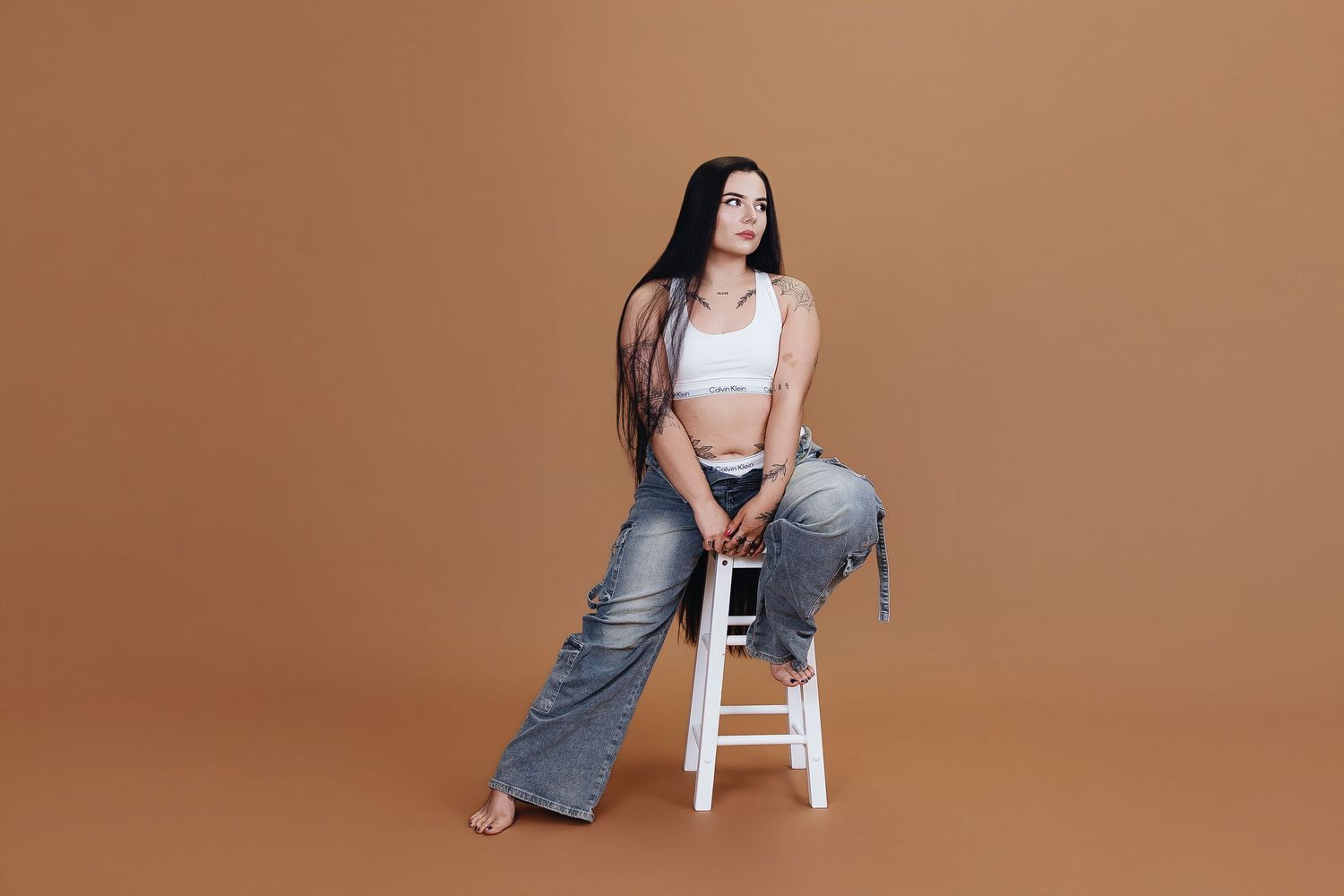 Young woman with long hair sits on white stool wearing white crop top and wide-leg jeans on brown background.