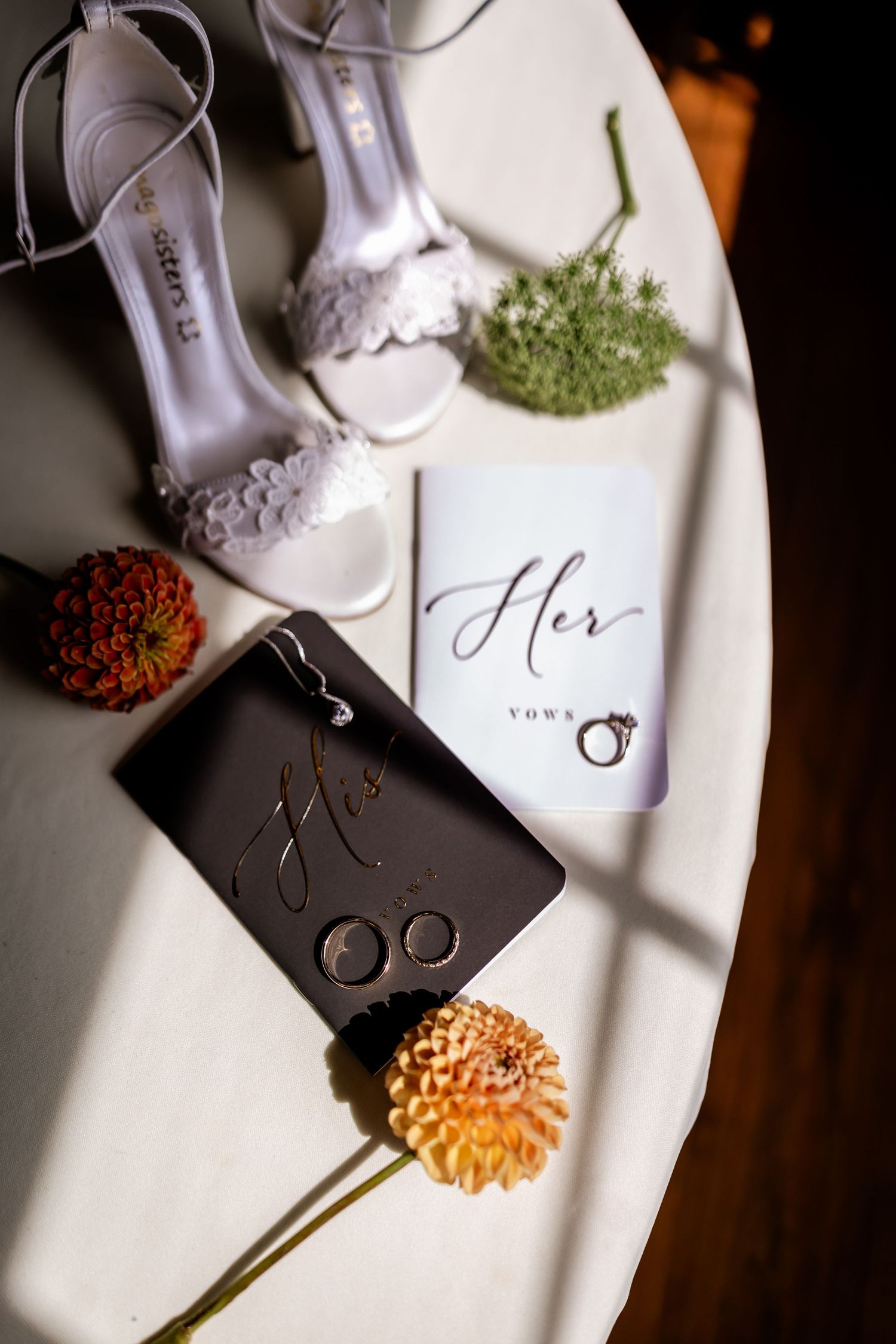Wedding flat lay with white heels, vow books, rings, and orange dahlias on a white table.