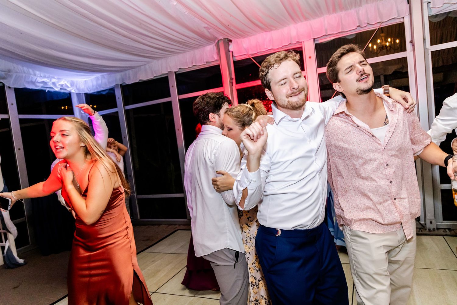 Wedding guests enjoying themselves on a dance floor under pink lighting and white draping.