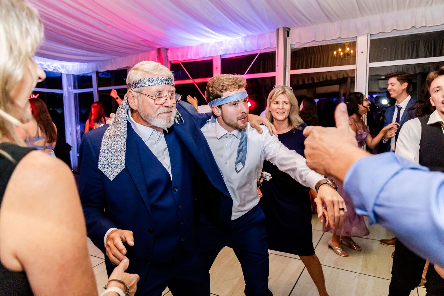 Wedding guests dancing and celebrating at reception under draped tent with colored lighting.