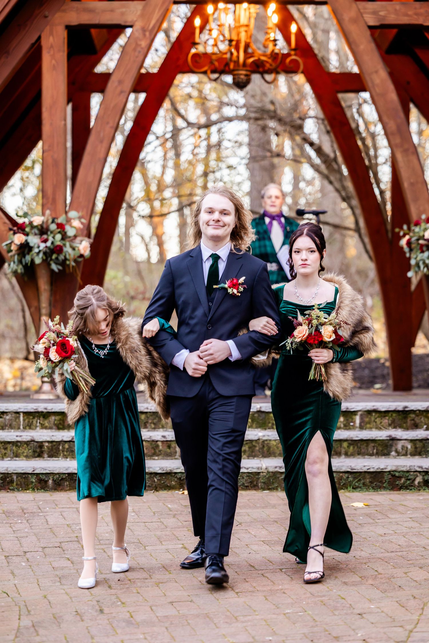 Wedding party members in elegant green dresses and navy suits walking down brick steps under a wooden archway.