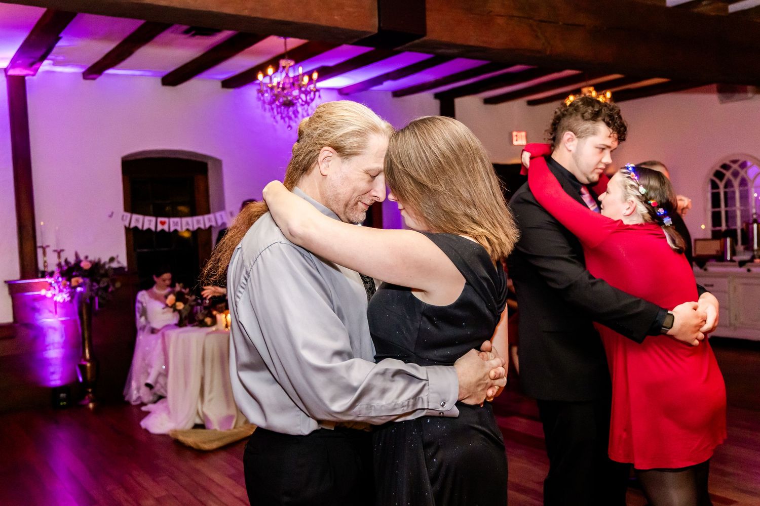 Wedding guests slow dancing in pairs on a dance floor illuminated by purple lighting and exposed wooden beams.