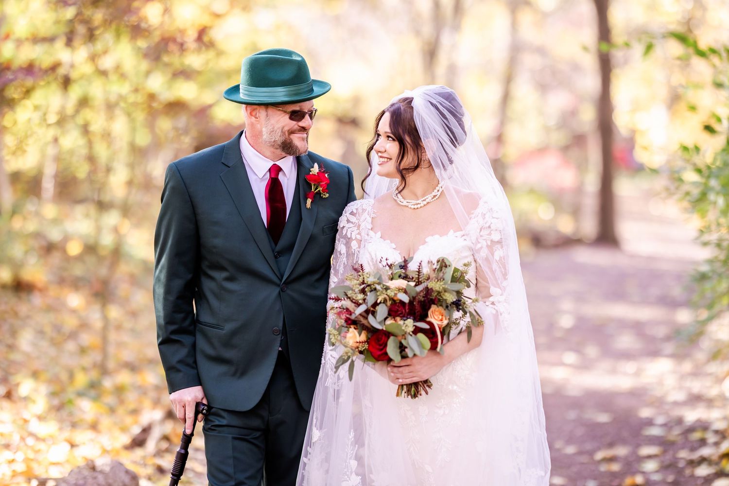 Newlywed couple walking together through a sunlit autumn forest path in formal wedding attire.