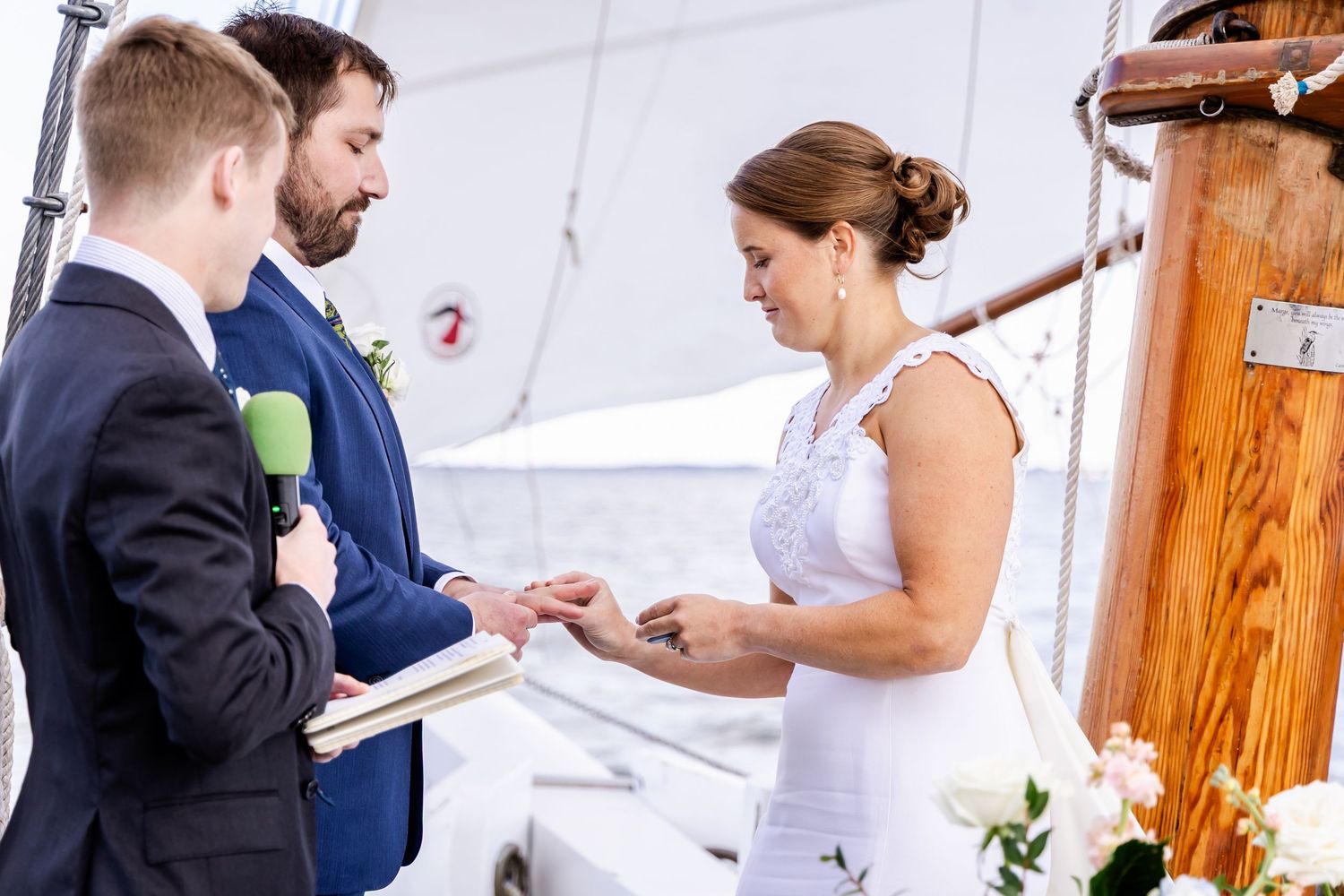 Couple exchanging wedding rings on a sailboat deck, officiated by a young man holding a microphone and book.