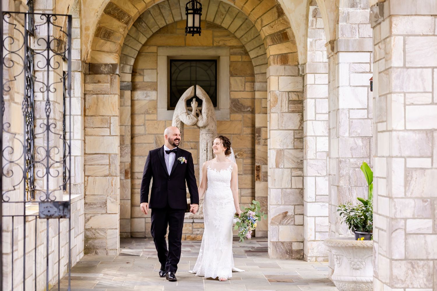 A bride and groom walk through a stone archway corridor with natural lighting filtering through the columns.
