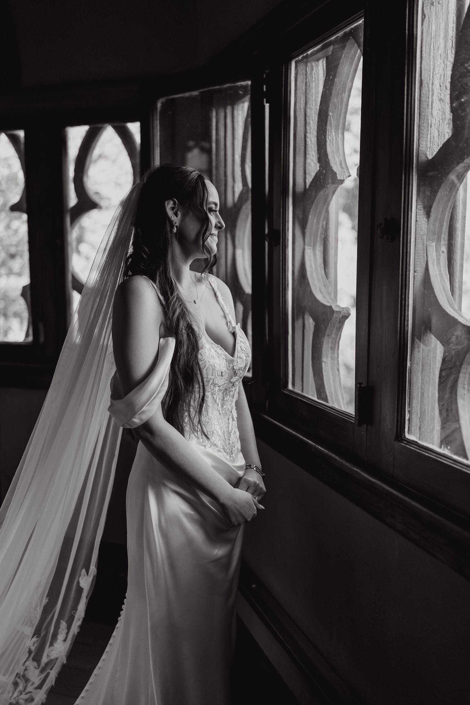 Black and white photograph of a bride in a wedding dress looking through a window in dramatic lighting.