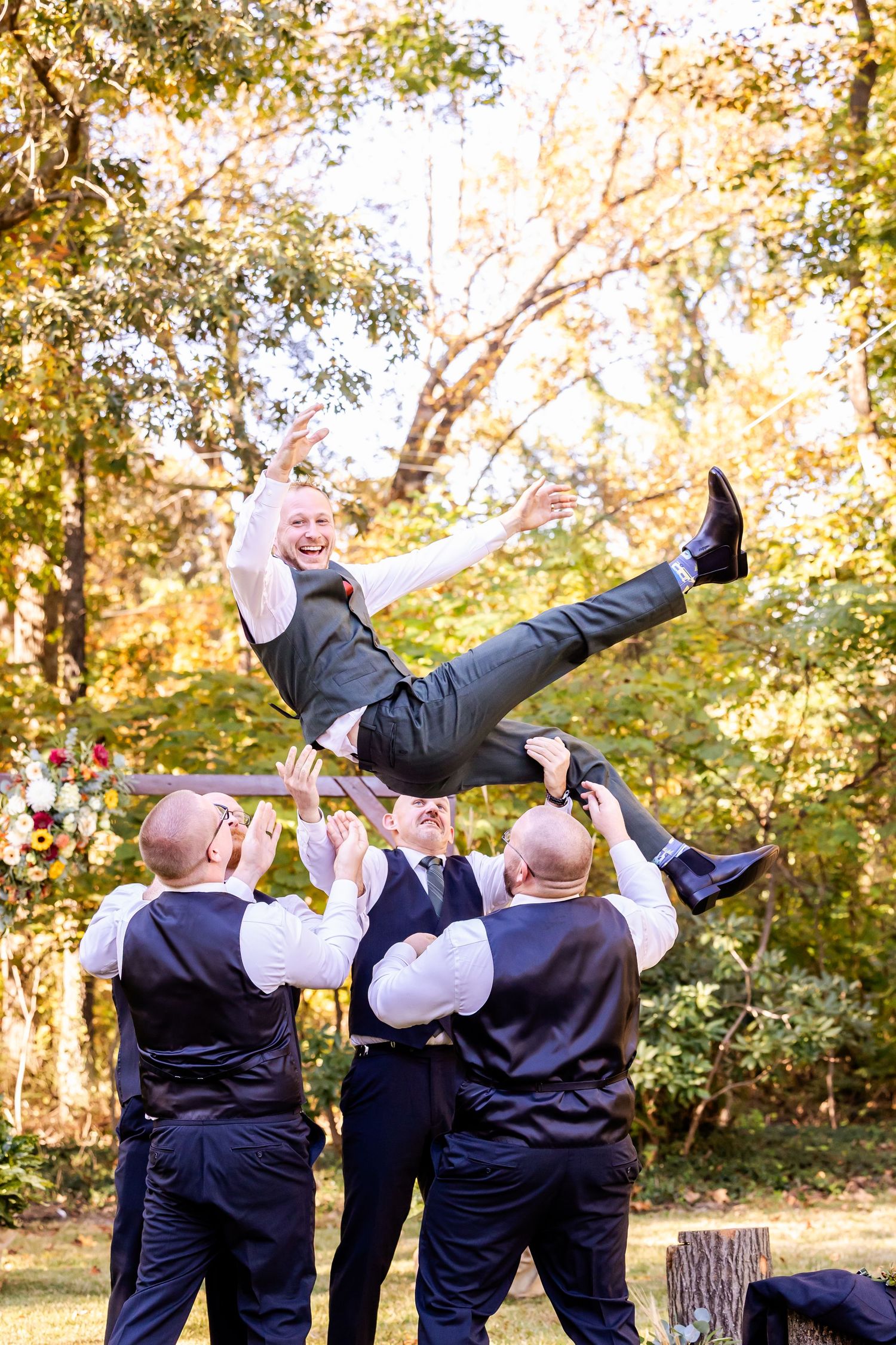 Groomsmen in navy suits playfully lift and toss their friend in the air during an outdoor autumn photoshoot.