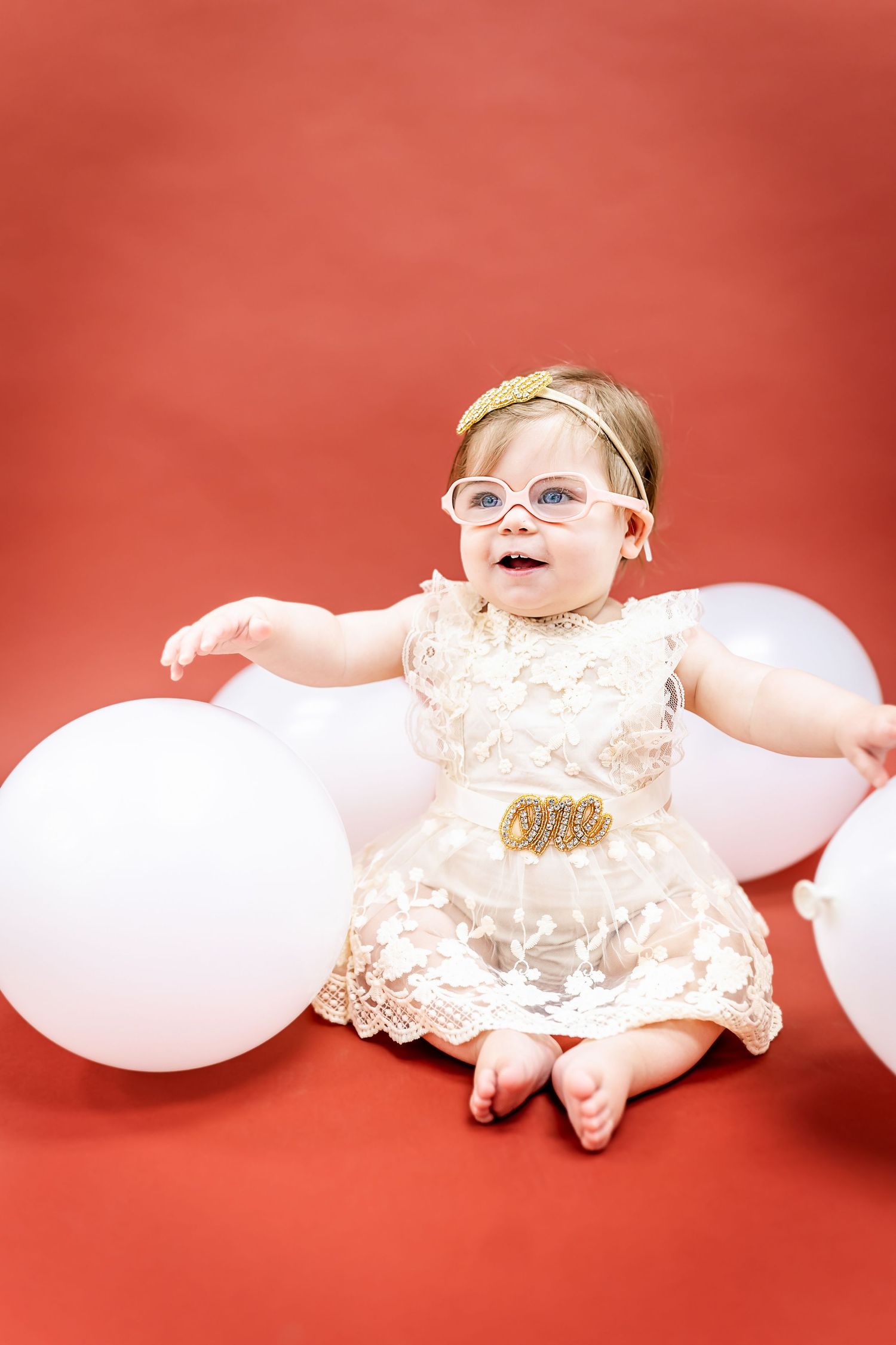 A baby in a lace dress sits with white balloons against a vibrant red background.