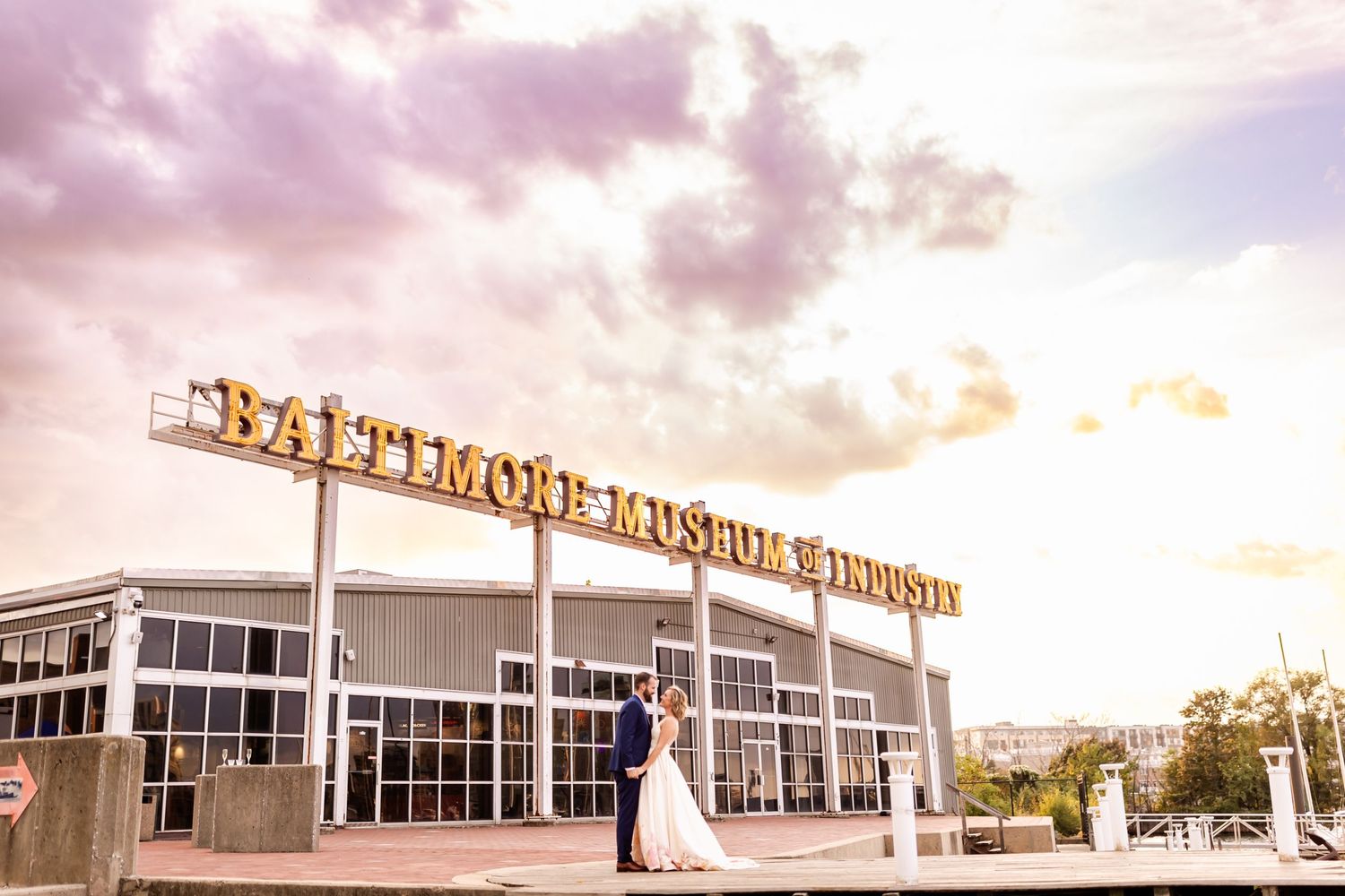 A couple embraces outside the Baltimore Museum of Industry building at sunset with dramatic pink and orange clouds.