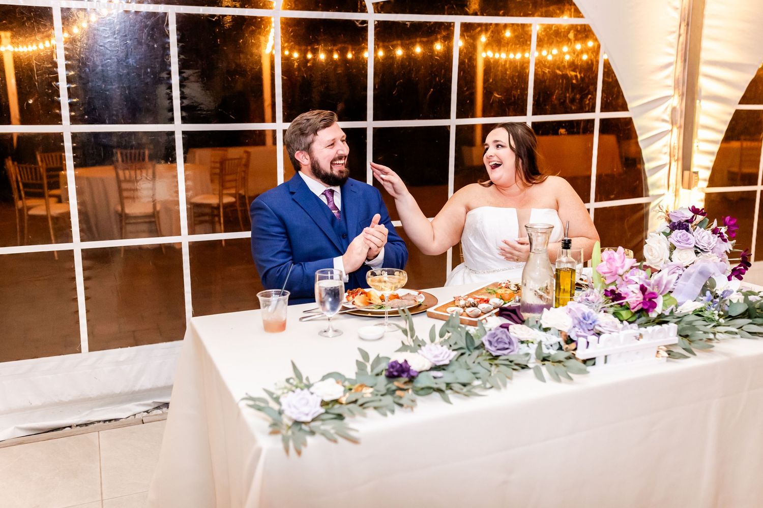 Couple sharing a toast at their wedding head table decorated with purple flowers and greenery.