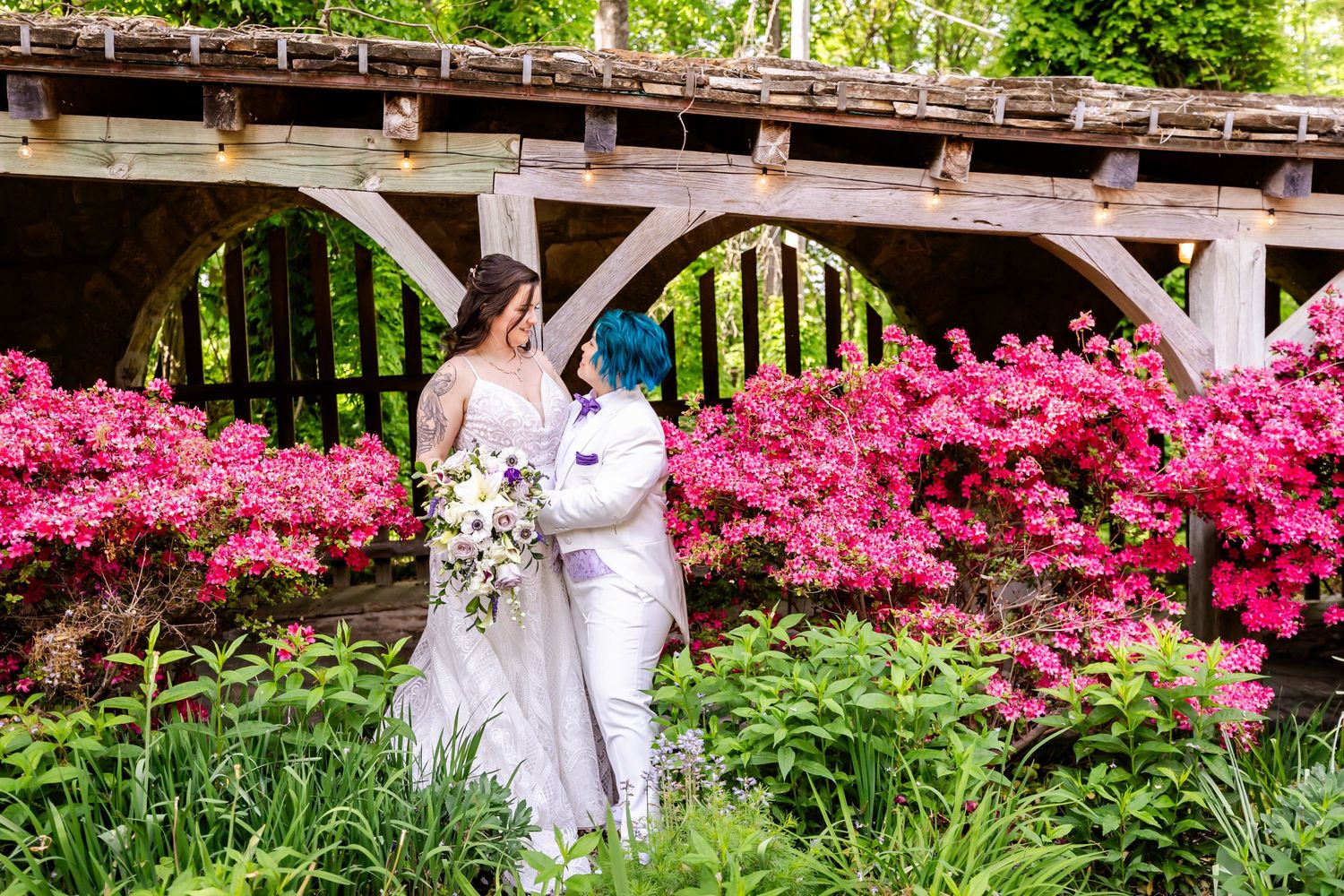 A wedding couple poses near vibrant pink azalea bushes under a rustic wooden pergola.