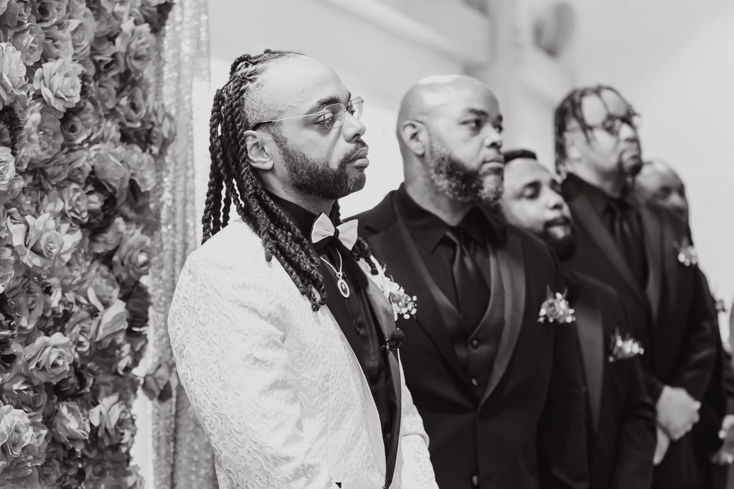 Groomsmen stand in line during wedding ceremony with floral backdrop in black and white.