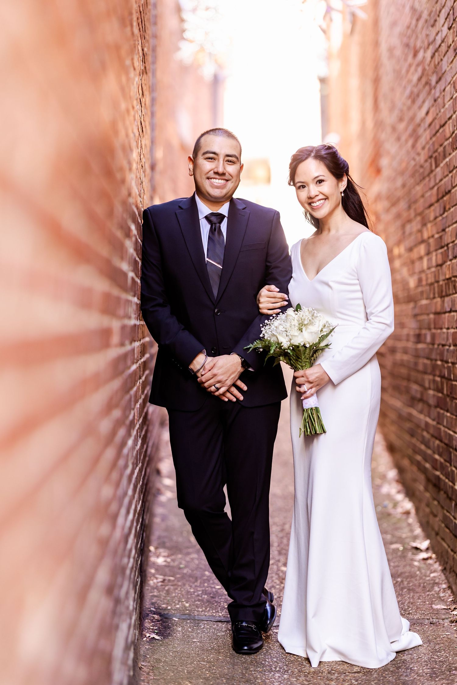 A wedding couple poses in a brick alleyway while the bride holds a white floral bouquet.