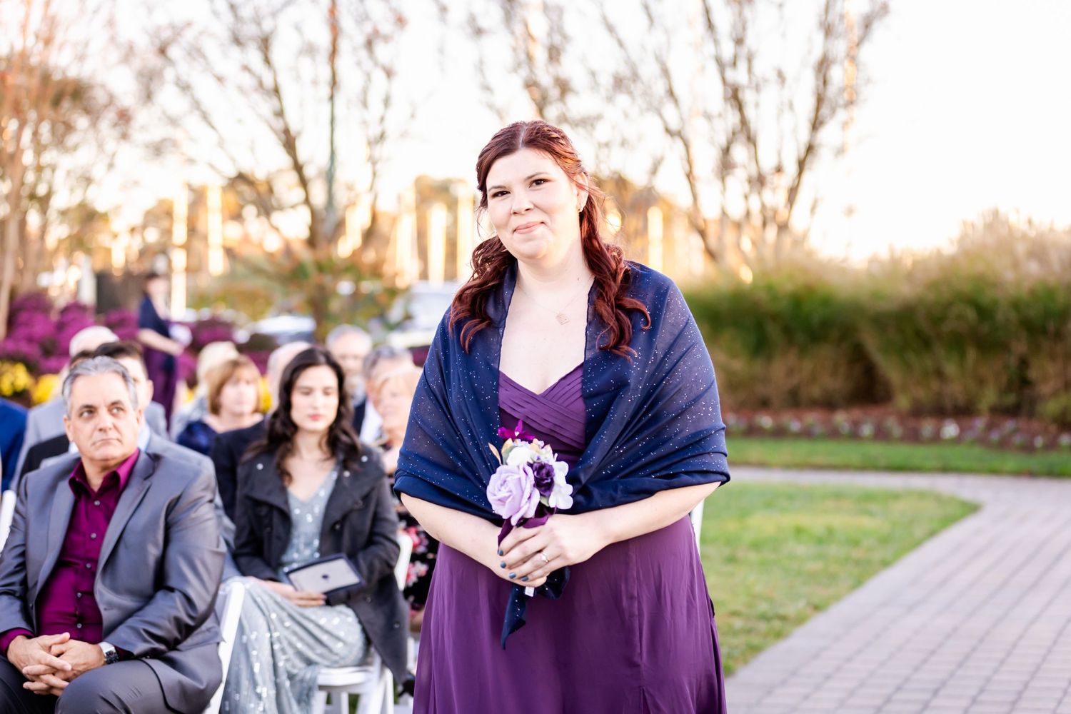 Wedding attendant in purple dress carrying flowers walks down aisle at outdoor ceremony.