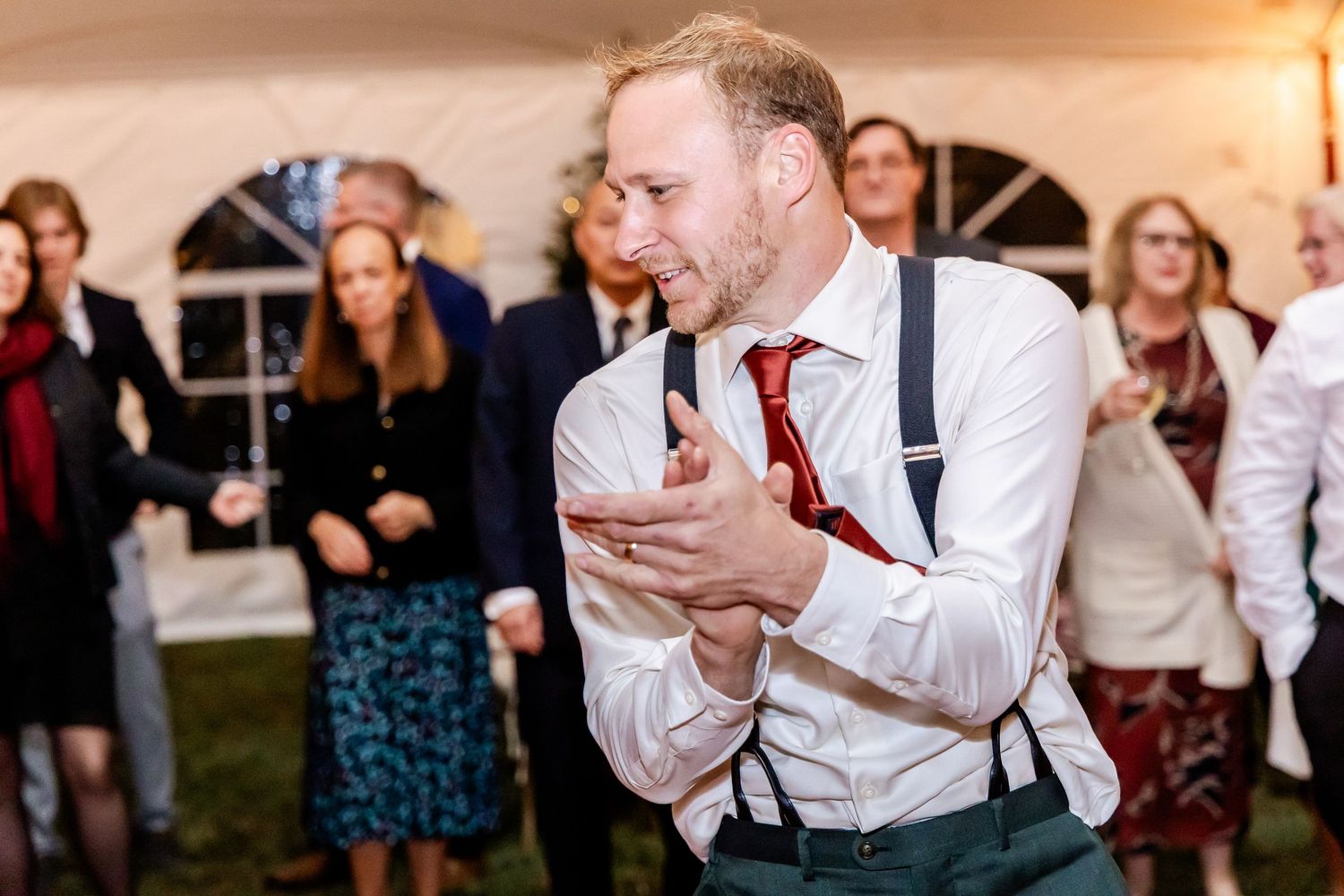 Wedding guests laugh and celebrate during an evening reception under a tent with string lights.